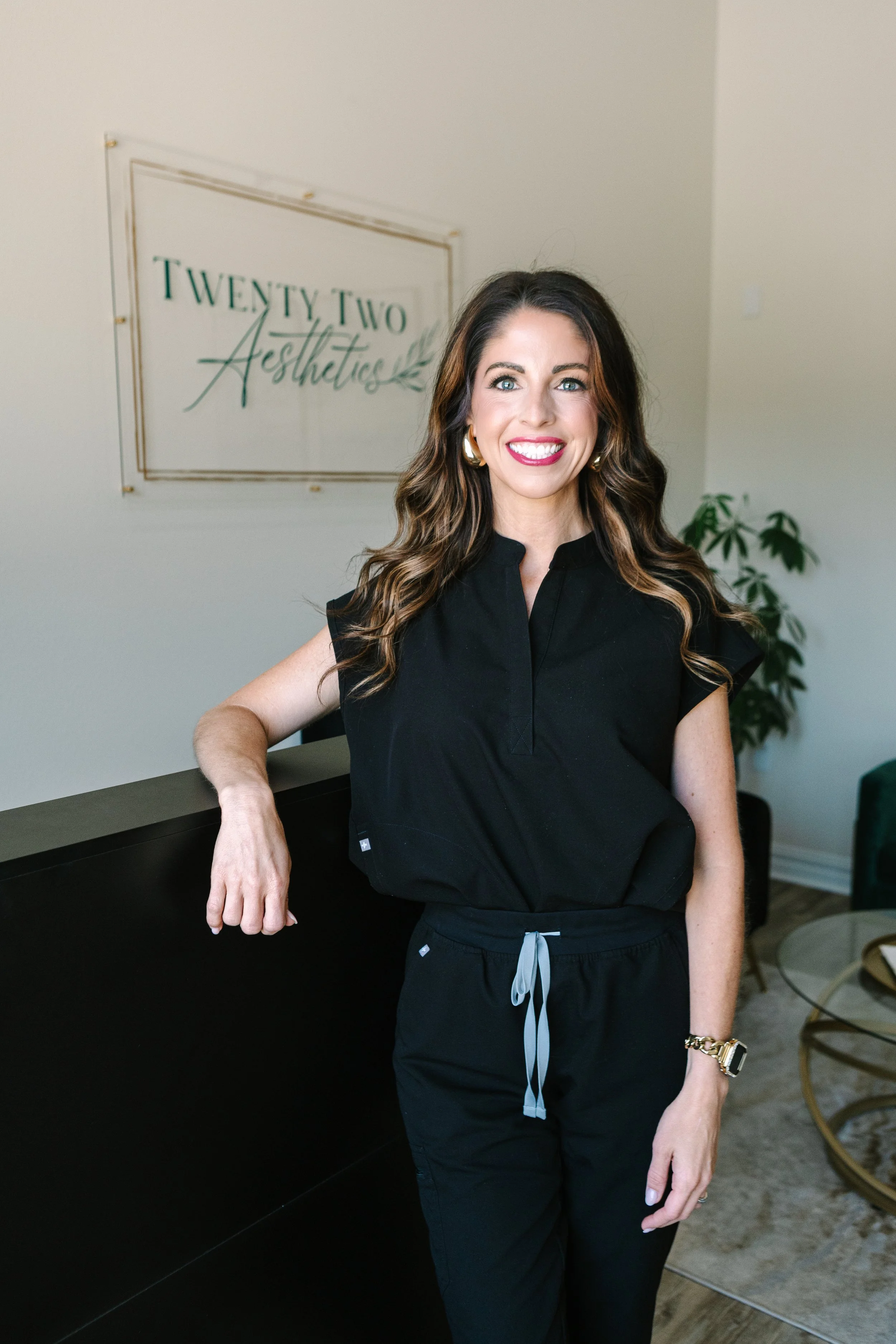 A woman with long brown hair, wearing a black sleeveless top and black pants, smiling and standing in an office or reception area with a sign that reads 'Twenty Two Aesthetics' in the background.