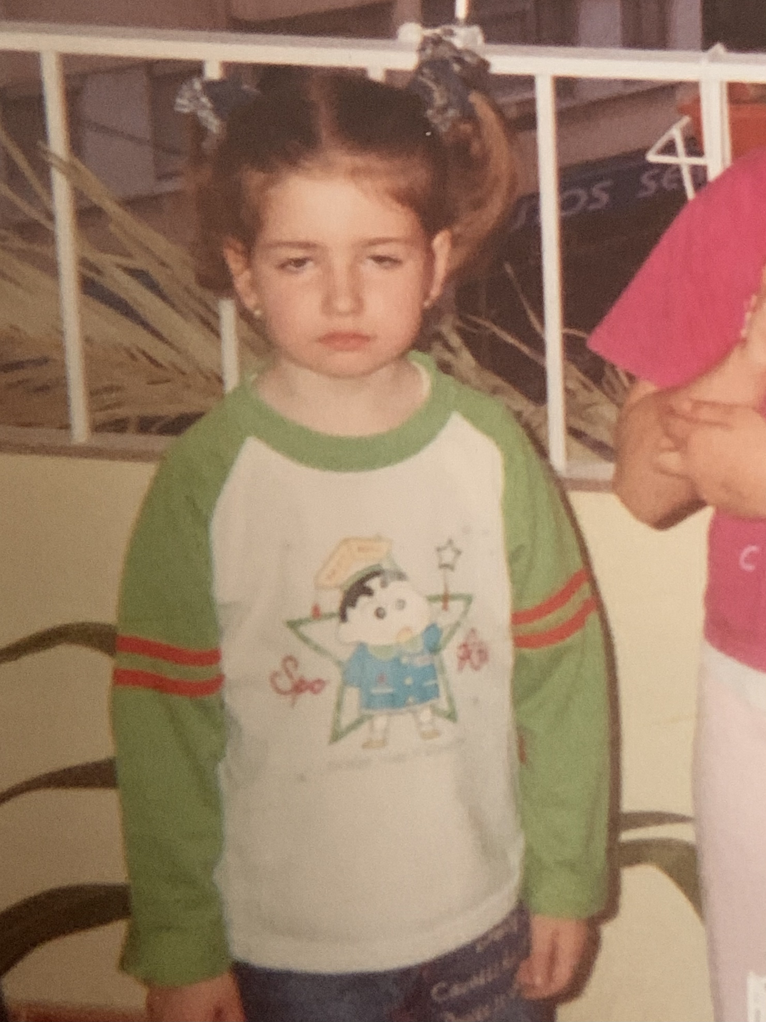 A young girl with red hair styled in pigtails, wearing a green and white shirt with cartoon character and star designs, standing with a neutral expression.