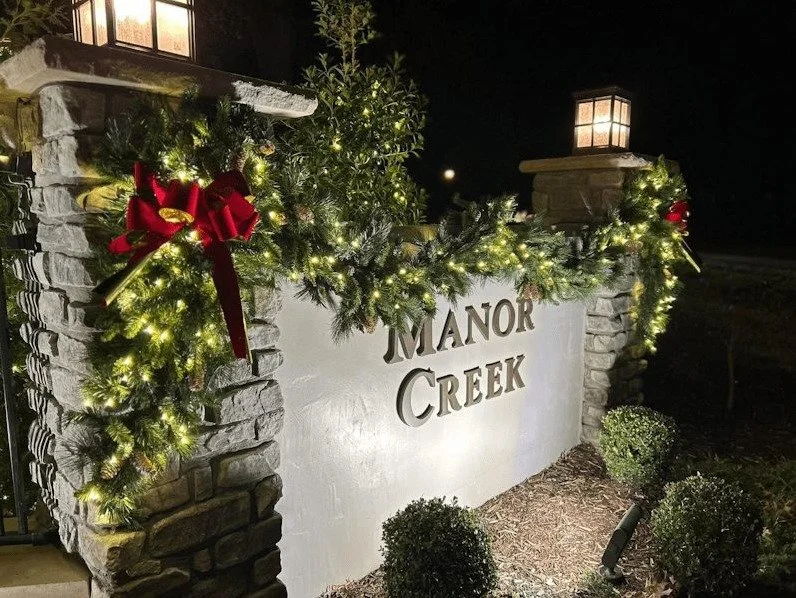 Decorated stone entrance sign with holiday lights, red bows, and greenery, with suburban houses and American flags in the background at sunset.