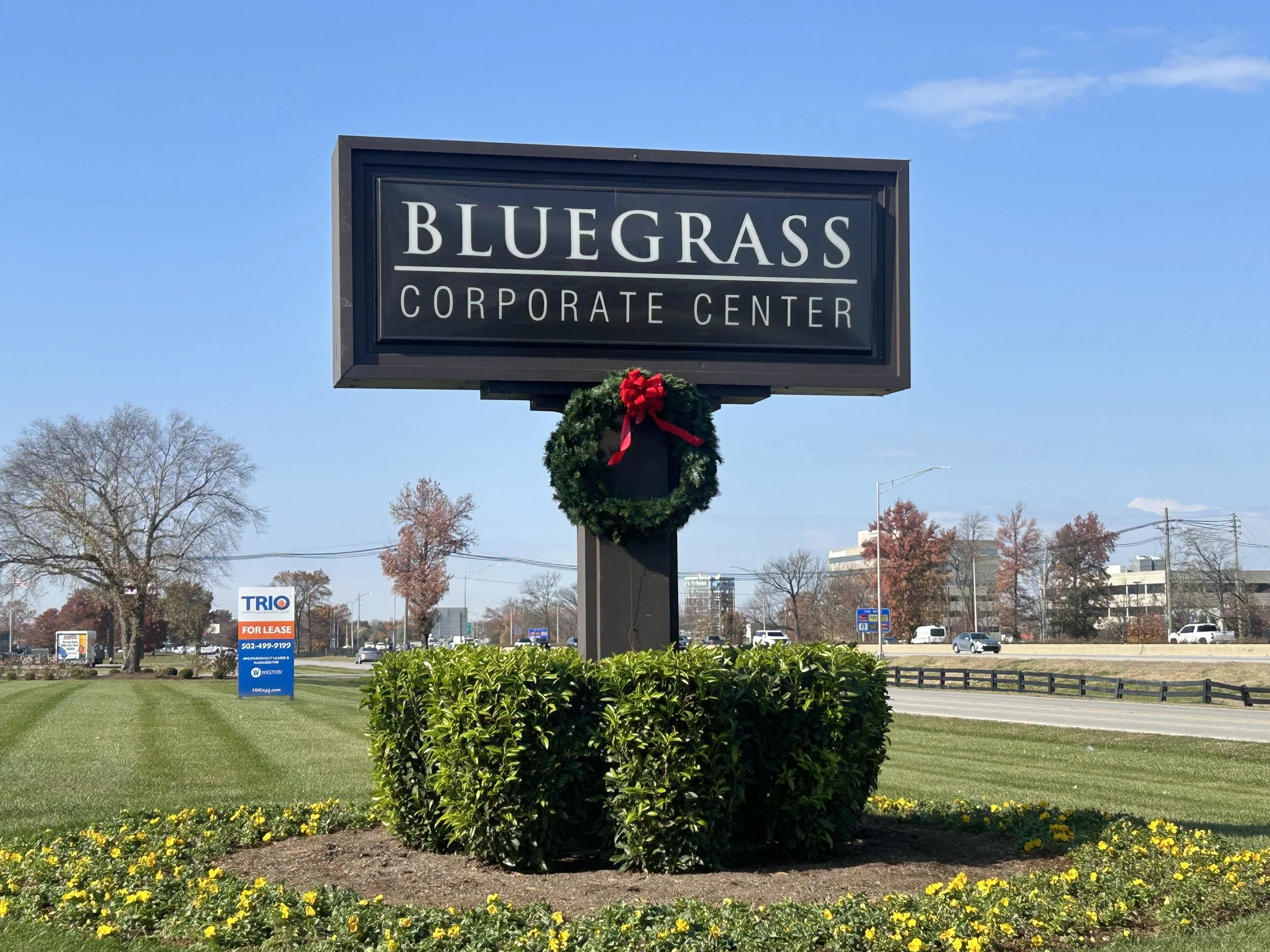 Sign for Bluegrass Corporate Center decorated with a holiday wreath, surrounded by bushes and grass, on a cloudy day with cars and buildings in the background.