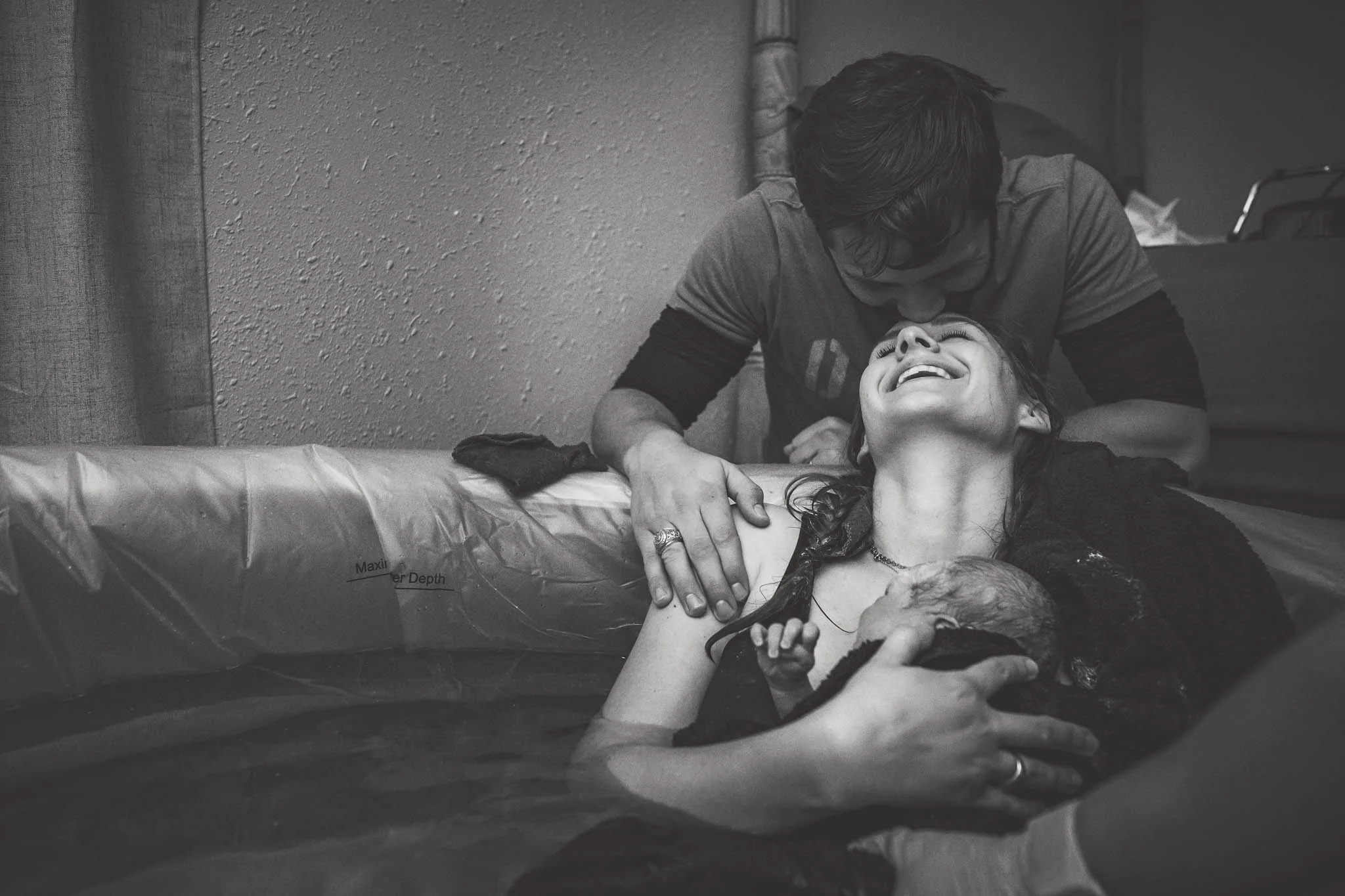 A woman in labor smiling and holding her newborn baby in a water birth in Cheyenne, with a man leaning over her in a supportive manner.