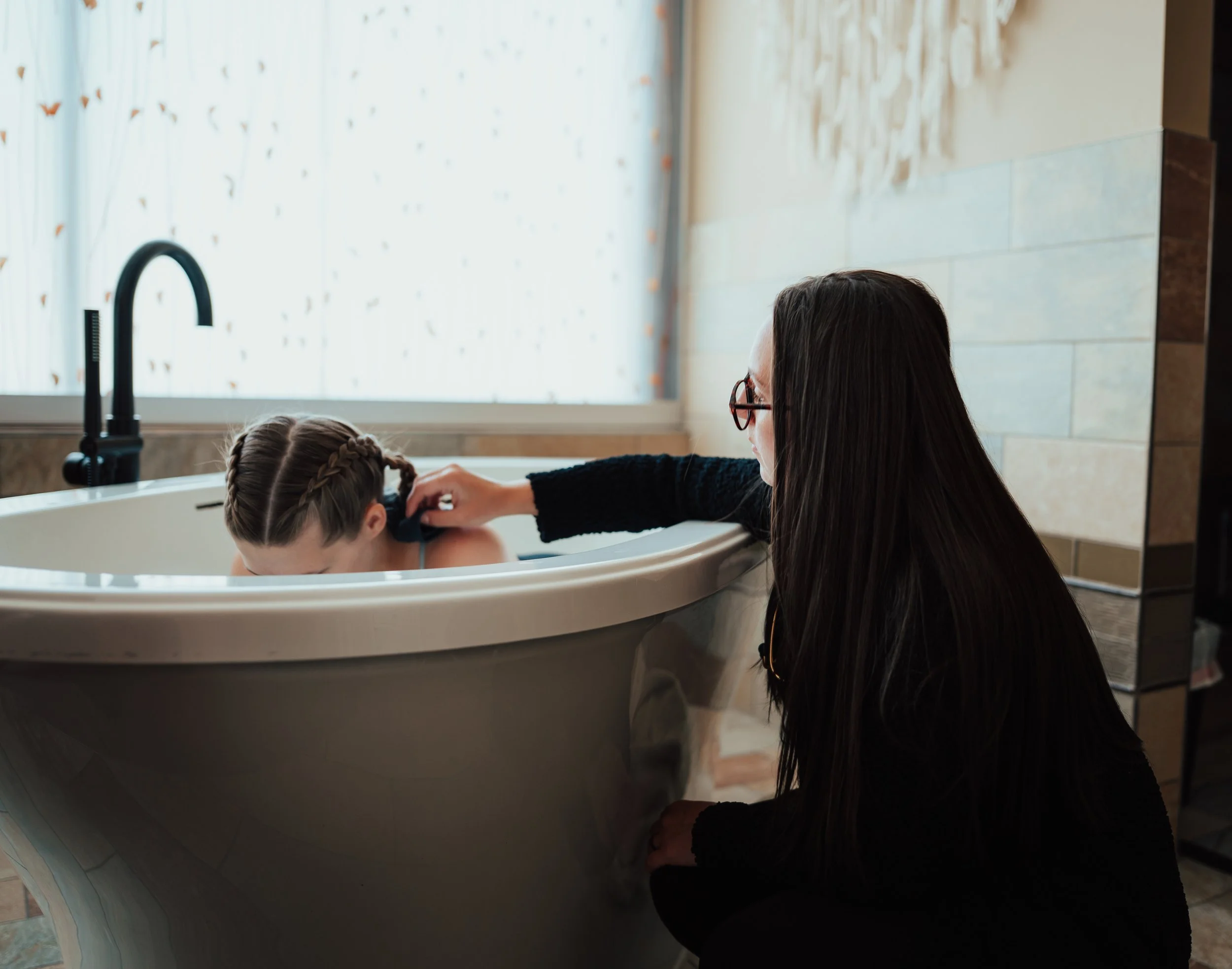 A woman with long dark hair wearing glasses and a black sweater is helping a young girl with braided hair get into a bathtub, which is in a well-lit bathroom with a window covered by sheer curtains.