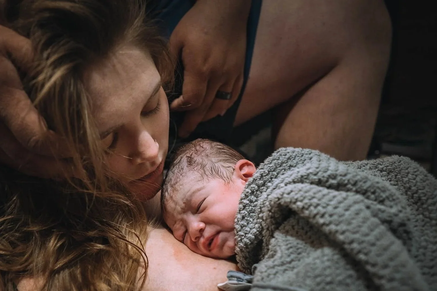 A woman with red hair and a nose ring cradles her crying newborn baby wrapped in a gray blanket, while a man supports her from behind, all in a tender, intimate moment.