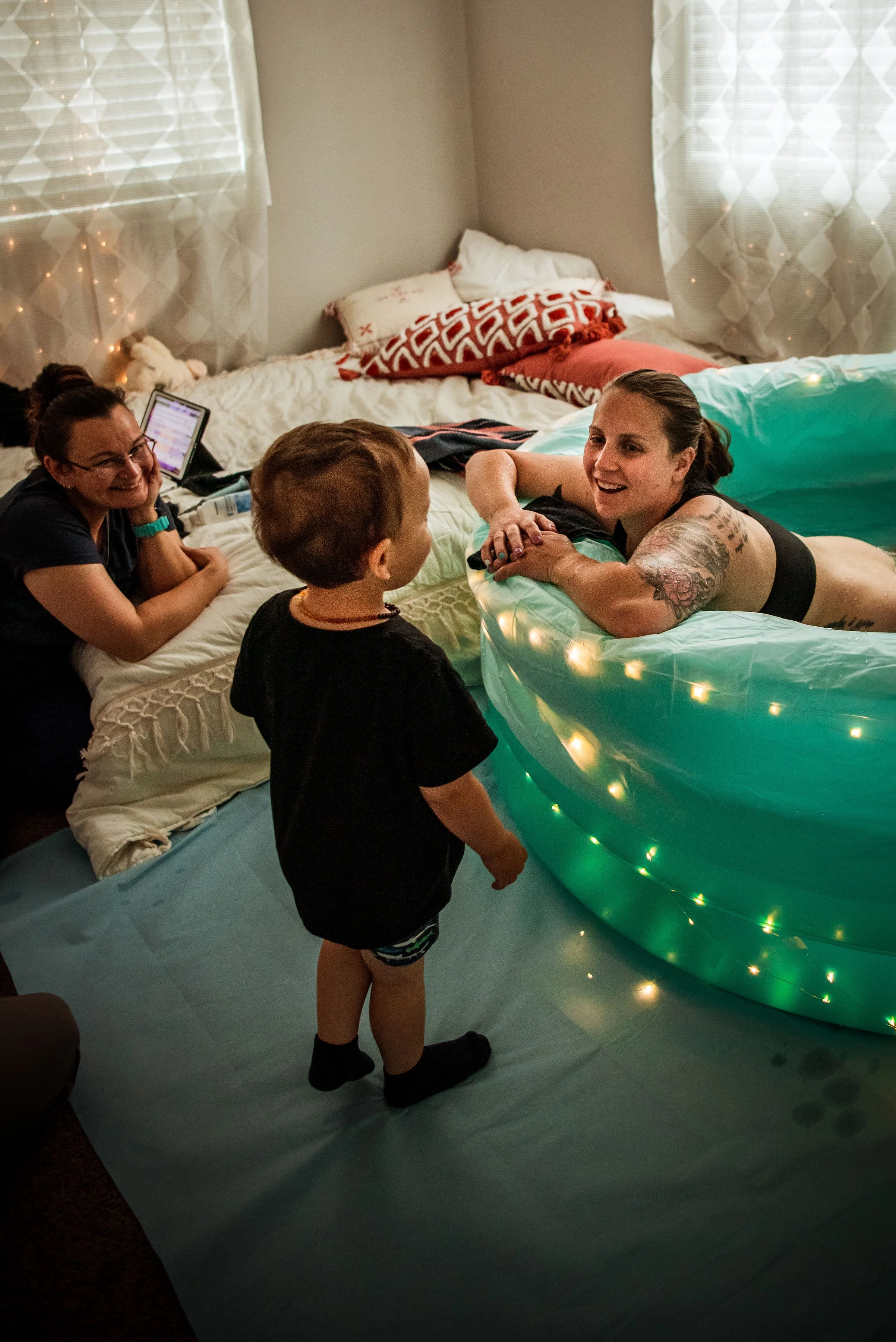 A woman with tattoos lying in an inflatable pool decorated with string lights, talking to a young boy standing nearby; another woman and a young girl are smiling and watching the interaction in a bedroom with bed, pillows, and fairy lights.