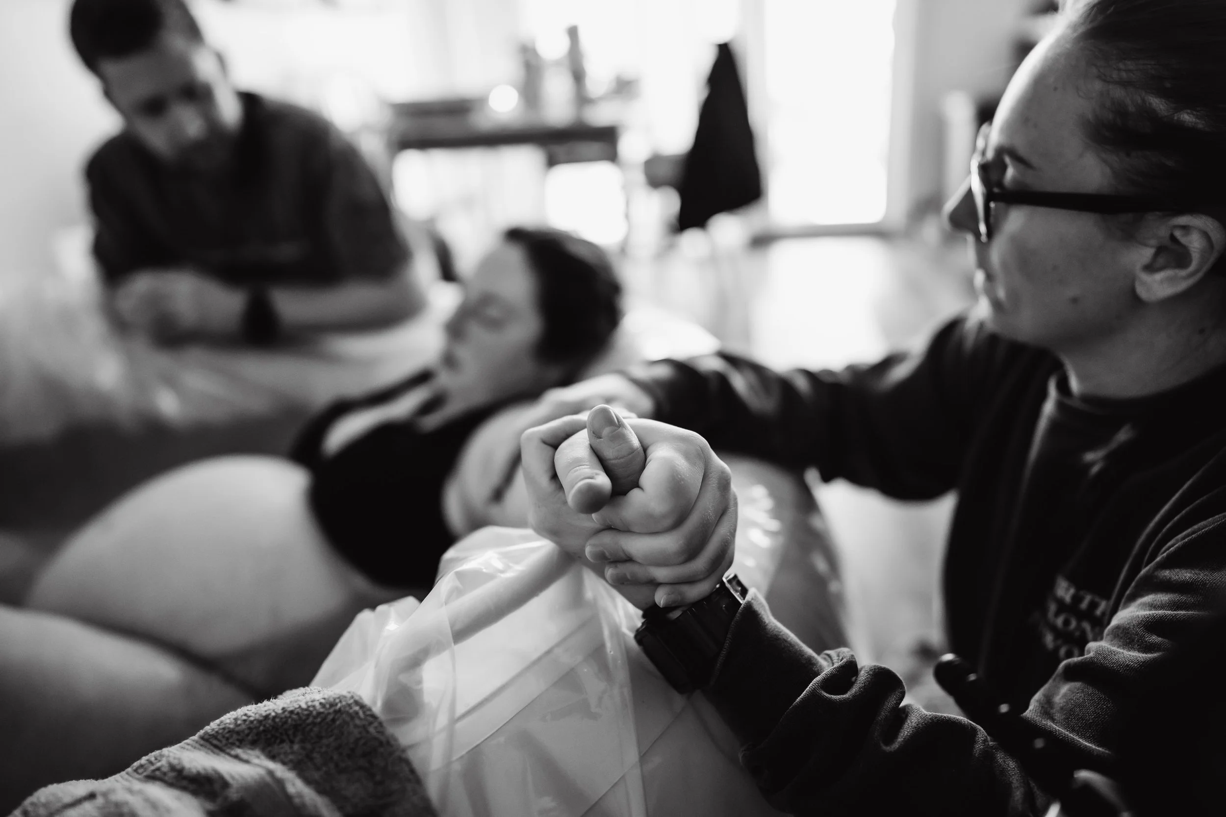 A woman lies on a hospital bed with her eyes closed while a man holds her hand, in a black-and-white photo.