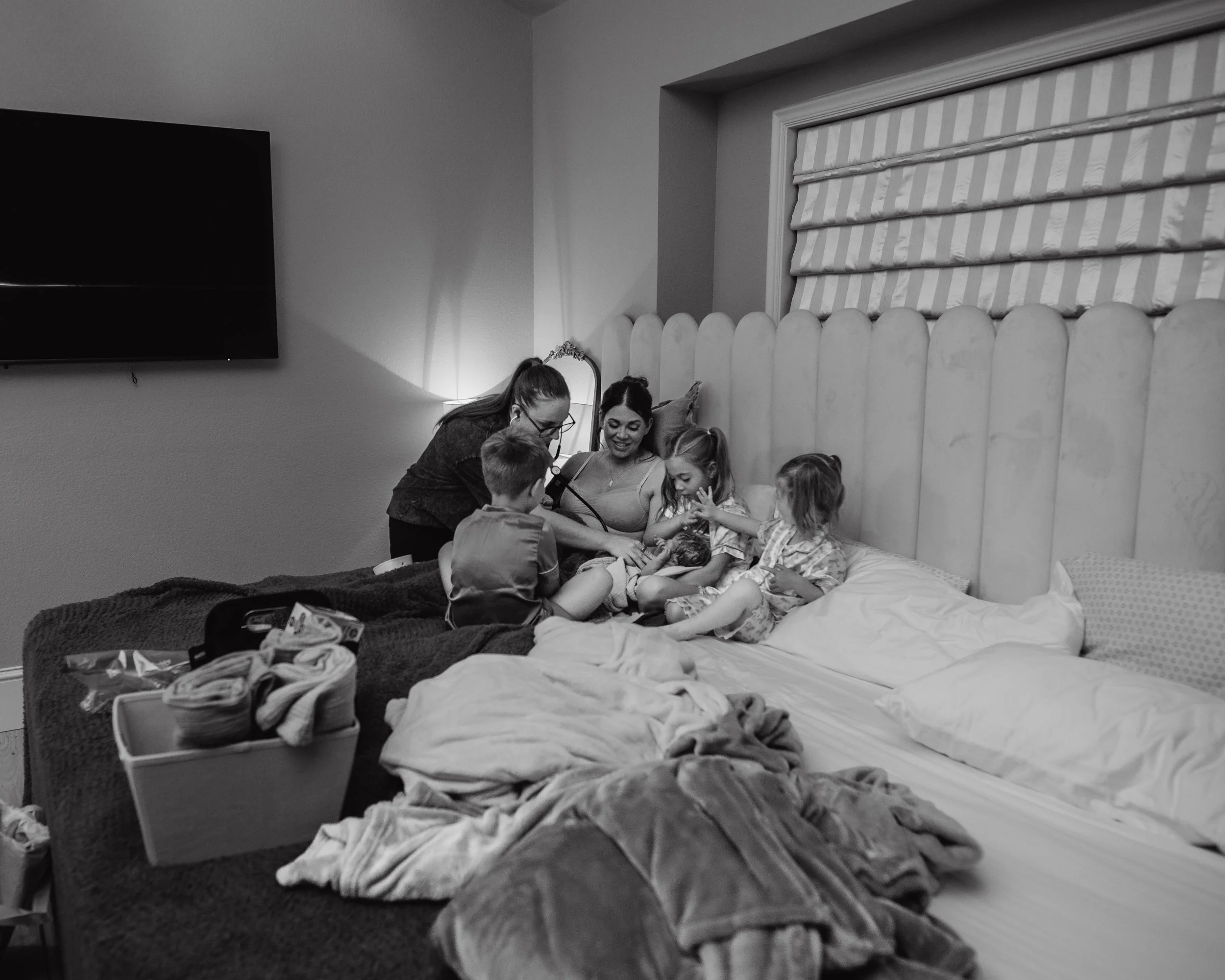 A woman and three young girls sitting on a bed with a nurse attending to a young girl with a stethoscope, in a hotel room setting.