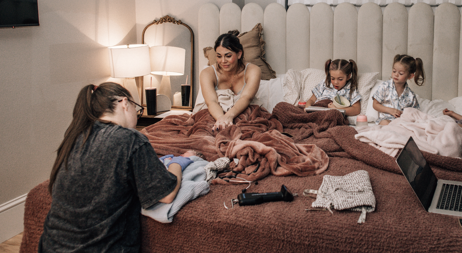 A woman and three young girls sit on a bed with blankets, engaging in different activities. The woman is leaning forward, smiling, while one girl in glasses is holding a baby doll, and the other two girls are sitting with one reading a book and the other looking on. A laptop and a camera are on the bed, and a bedside table with lamps is in the background.