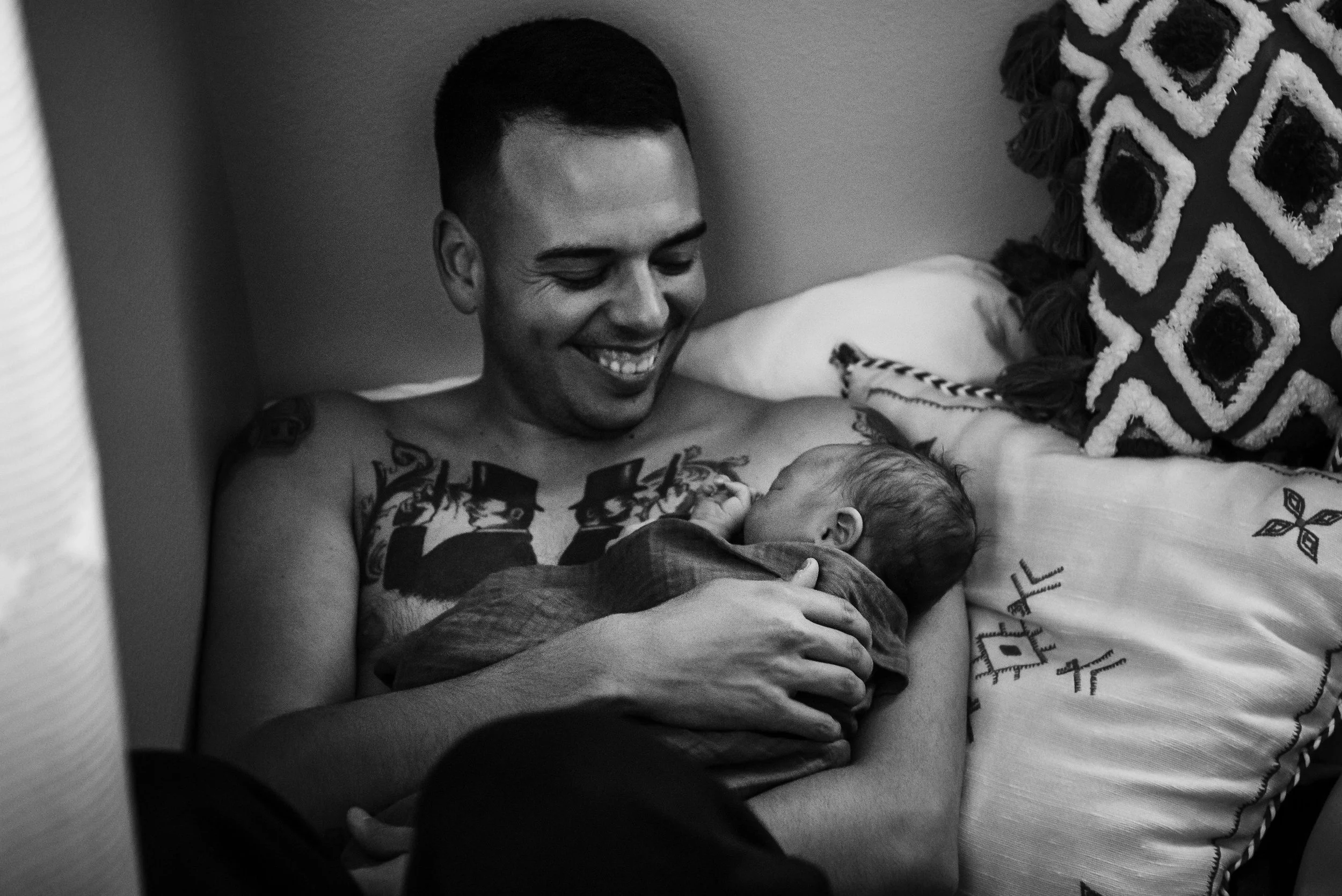 A smiling man holding a newborn baby in his arms while lying on a bed.