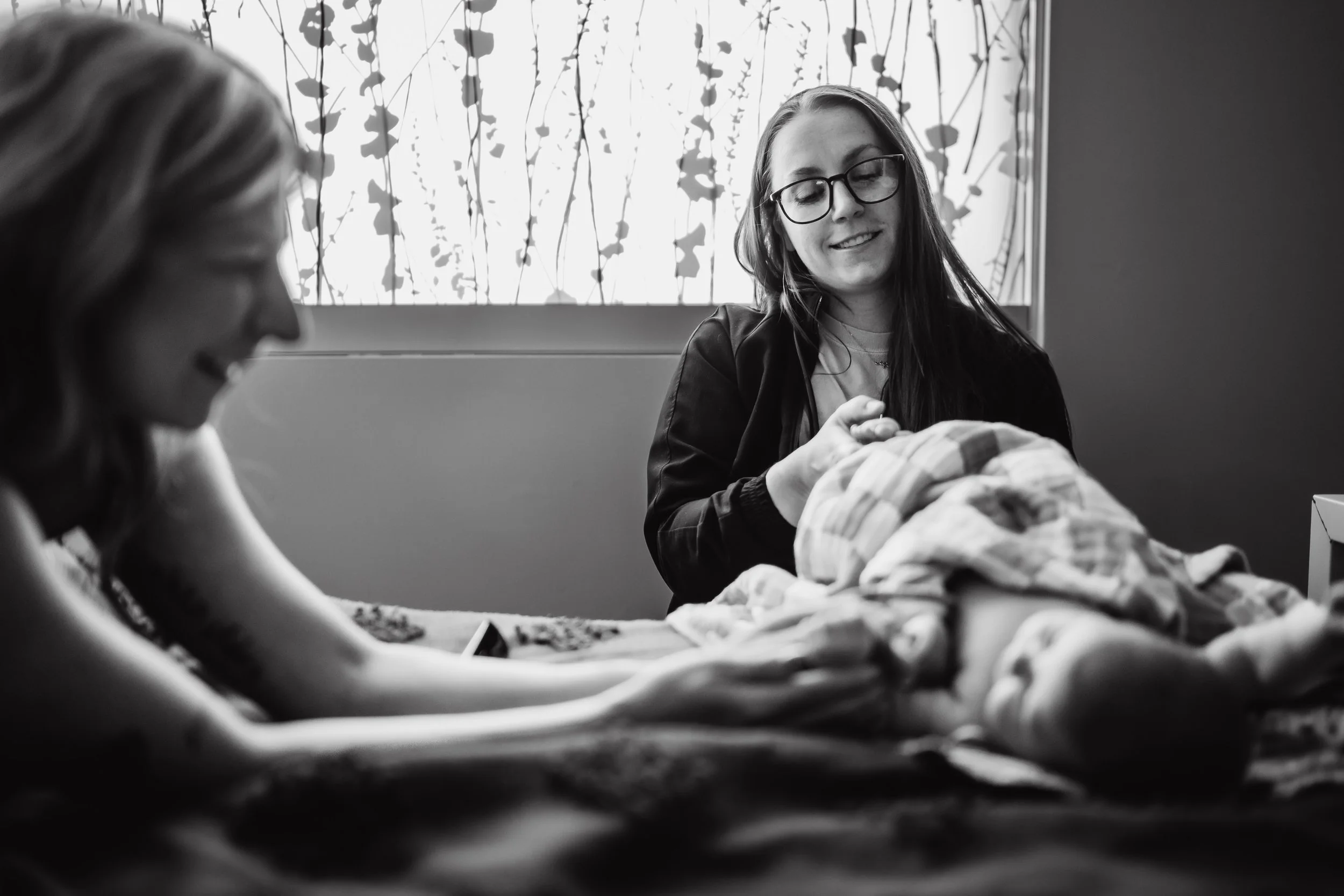 Two women with long hair are in a room with a window, one of them is holding a baby, and the other is observing. The baby is lying on a bed covered with a quilt. The women are smiling, and the woman with glasses is looking at the baby.