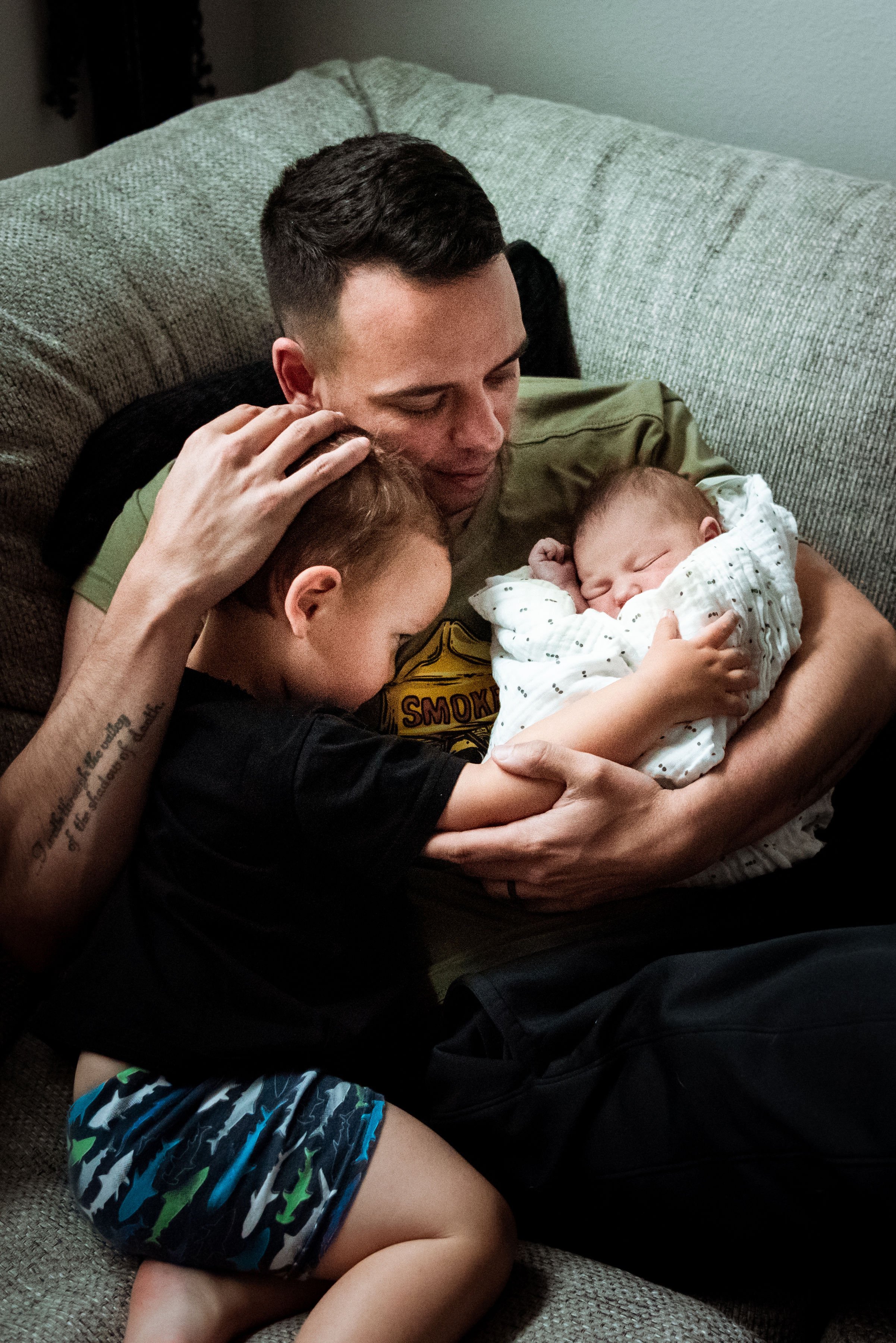 A man, two children, and a newborn baby cuddling on a gray sofa. The man gently holds the baby, while one child hugs the baby, and the other child rests on his shoulder, all appearing to share a tender moment.