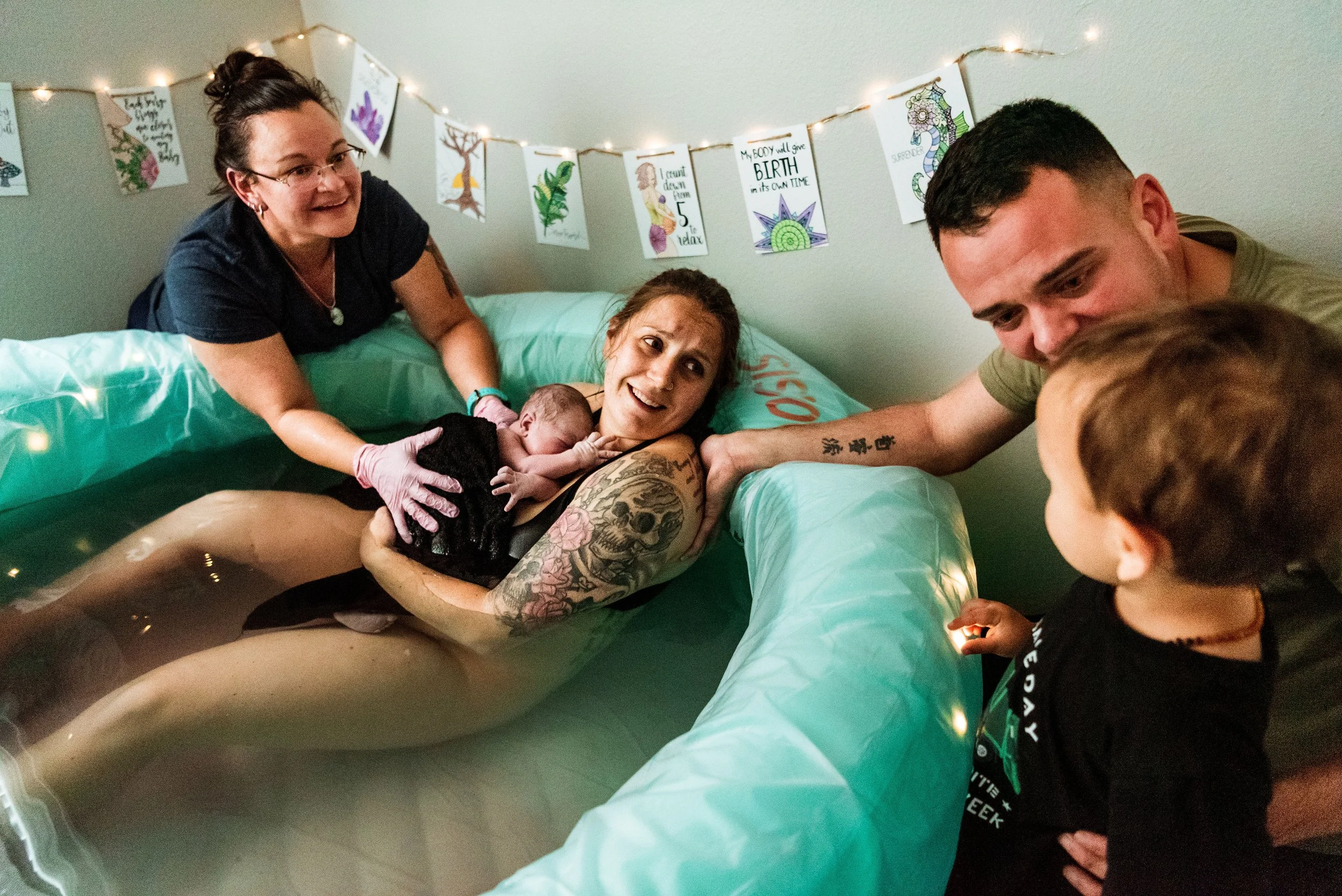 Woman giving birth in a birthing tub, surrounded by family members, including a child and two adults, with decorative lights and artwork in the background.