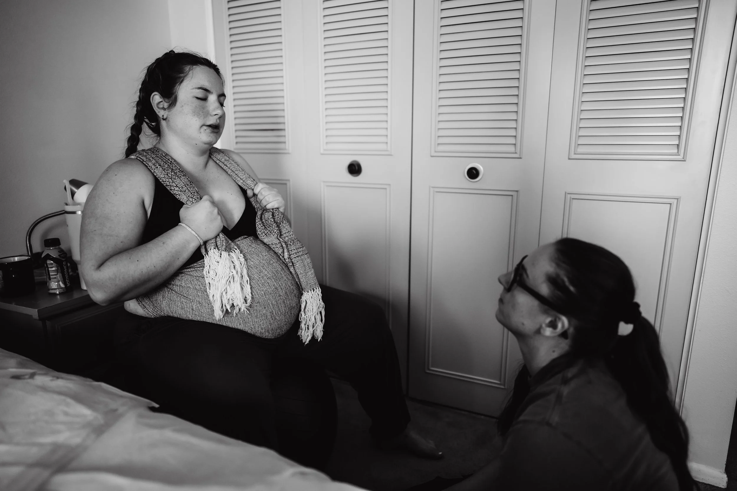A woman with a backpack sitting on a bed with her eyes closed, and another woman kneeling before her, looking up, in a bedroom with closed closet doors.