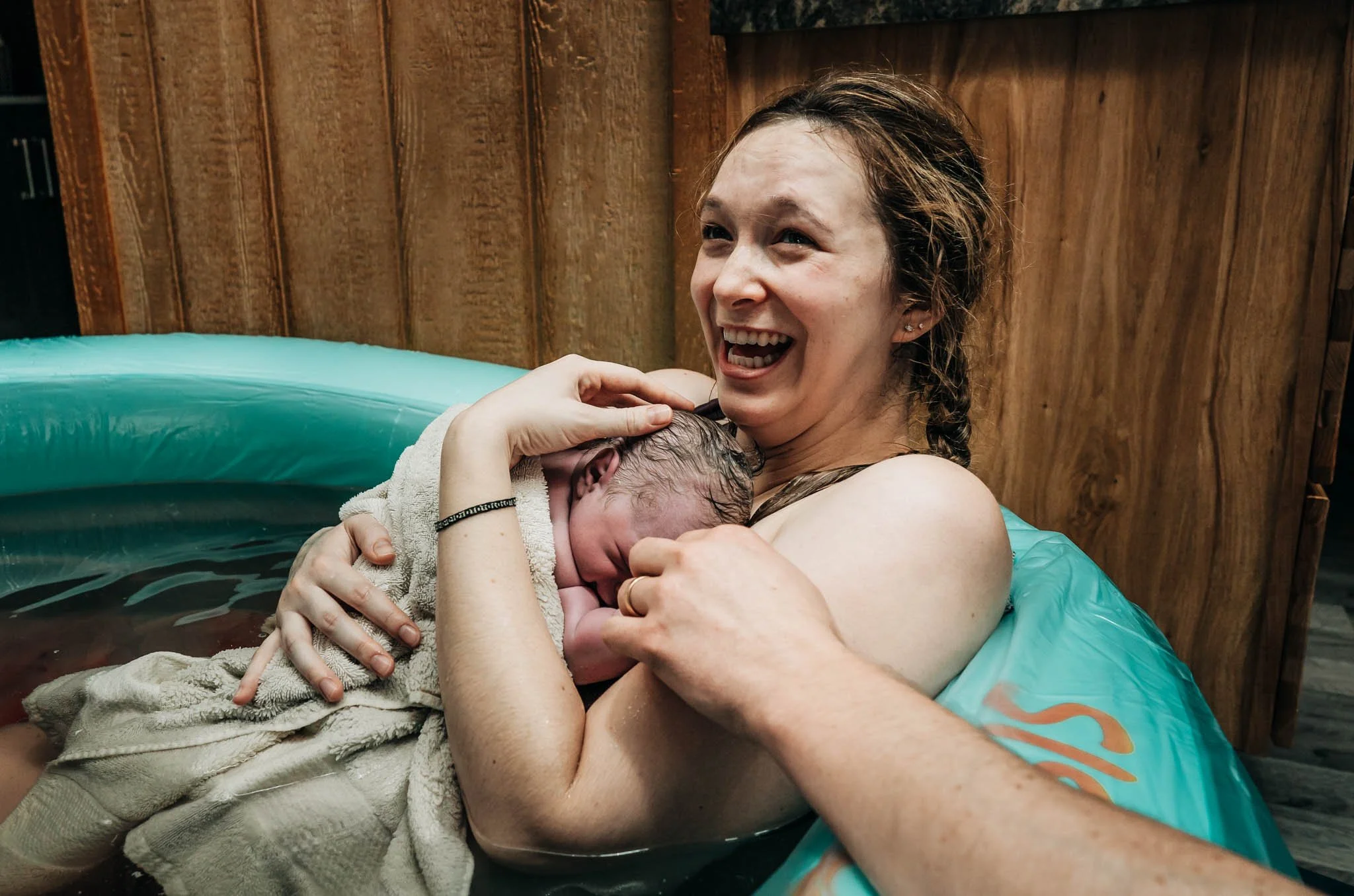 A woman happily giving birth in a birthing tub, holding her newborn baby close to her chest.
