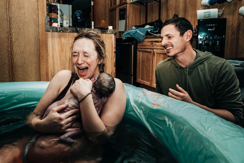 Woman holding a newborn baby in a small inflatable pool, smiling and crying; man beside the pool smiling and watching.
