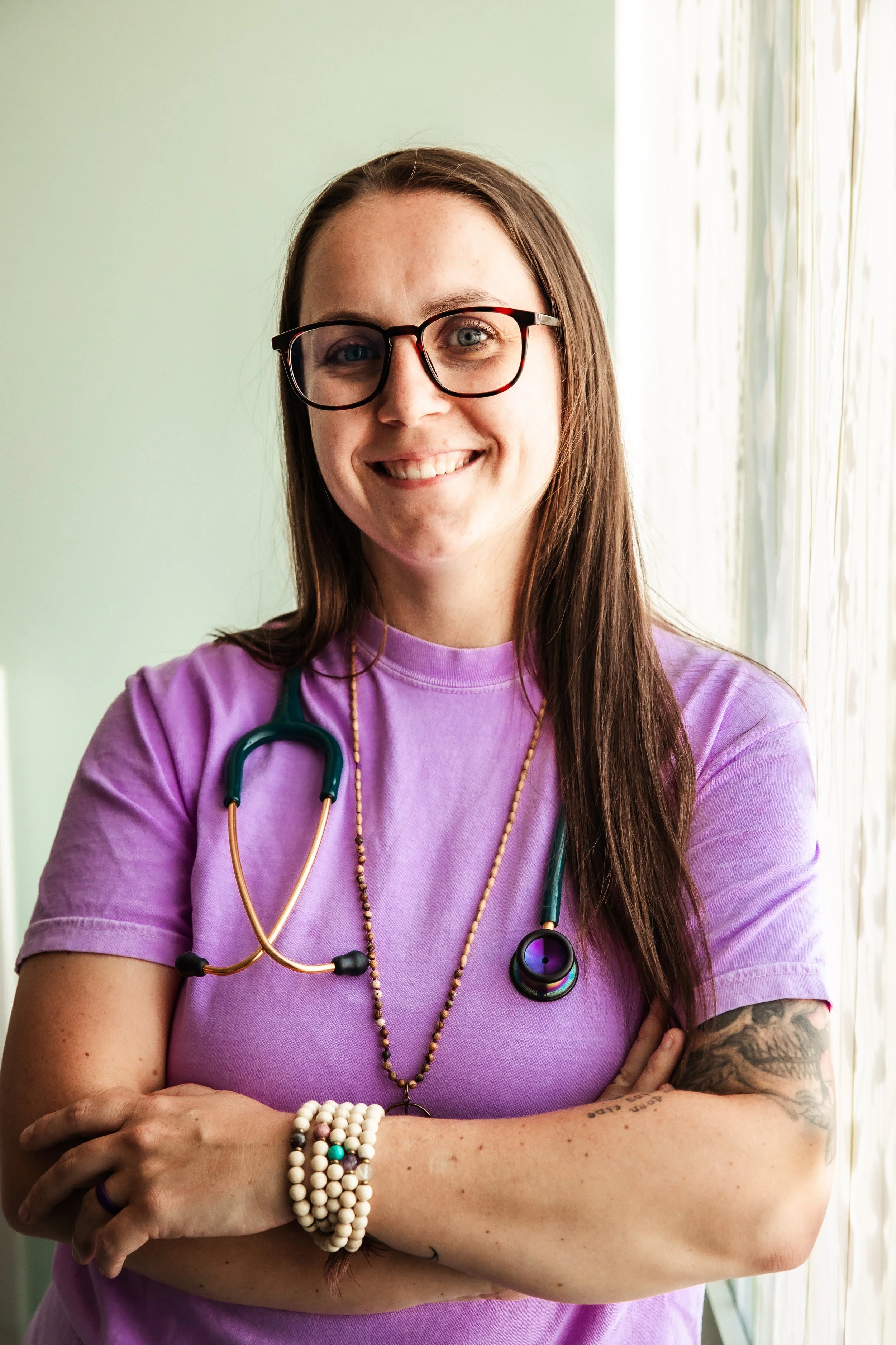 A smiling woman with long brown hair, glasses, and tattoos, wearing a purple shirt and a stethoscope around her neck, standing with crossed arms near a window with light-colored curtains.