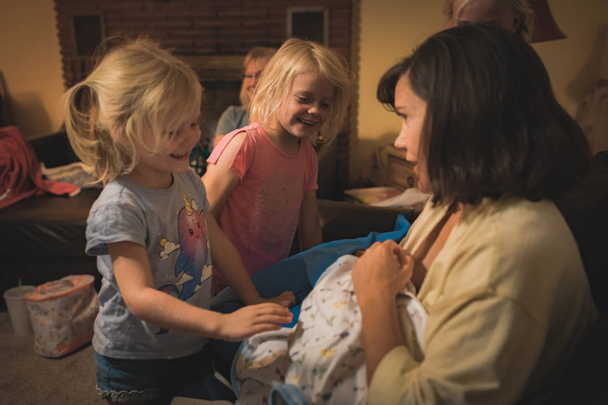 Two young girls and a woman with dark hair gather around a woman holding a newborn baby, in a cozy living room with a fireplace in the background. The girls are smiling and touching the baby.