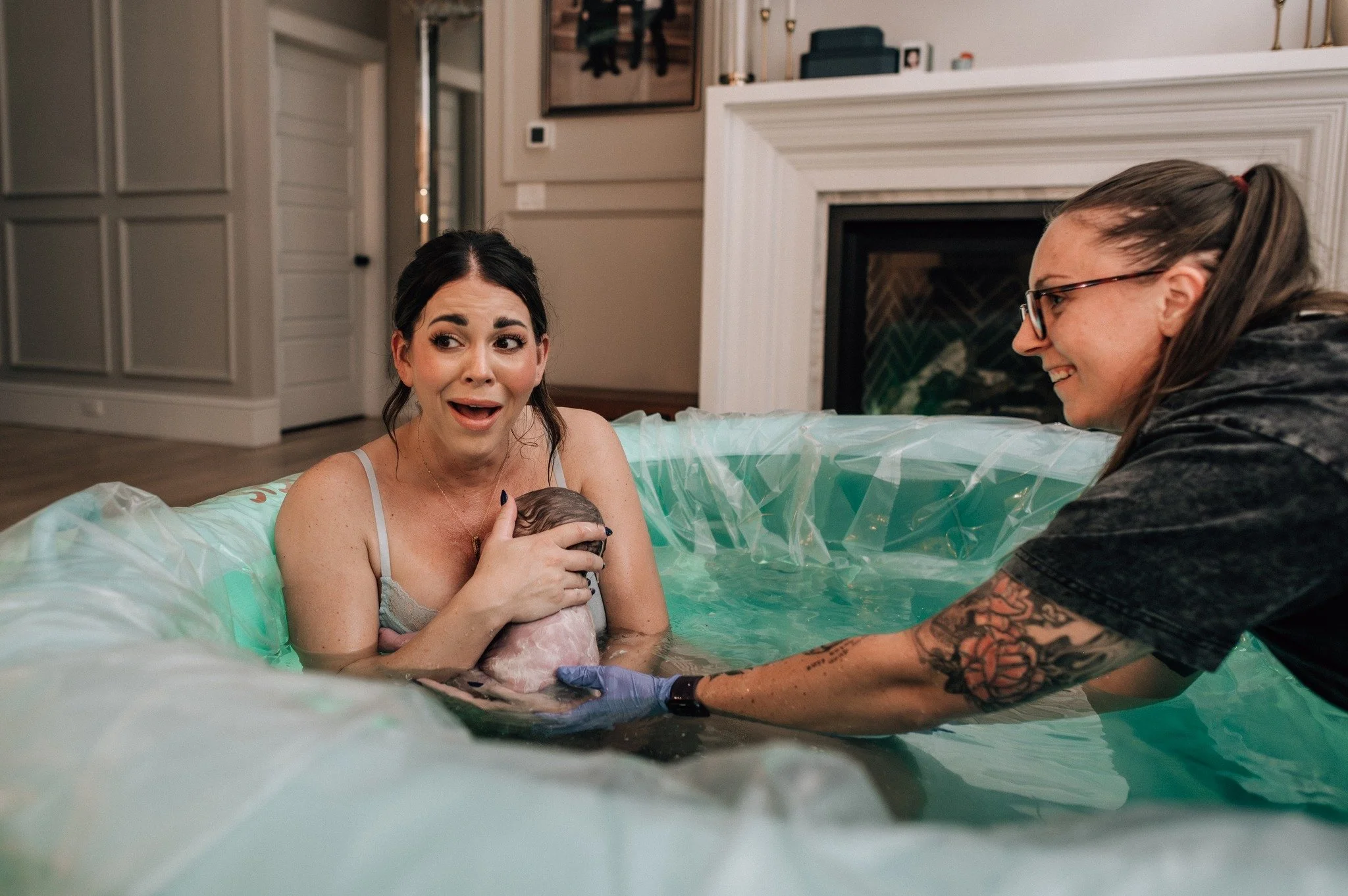 Woman giving birth in a birthing pool, with a midwife or partner supporting her and another woman smiling nearby.