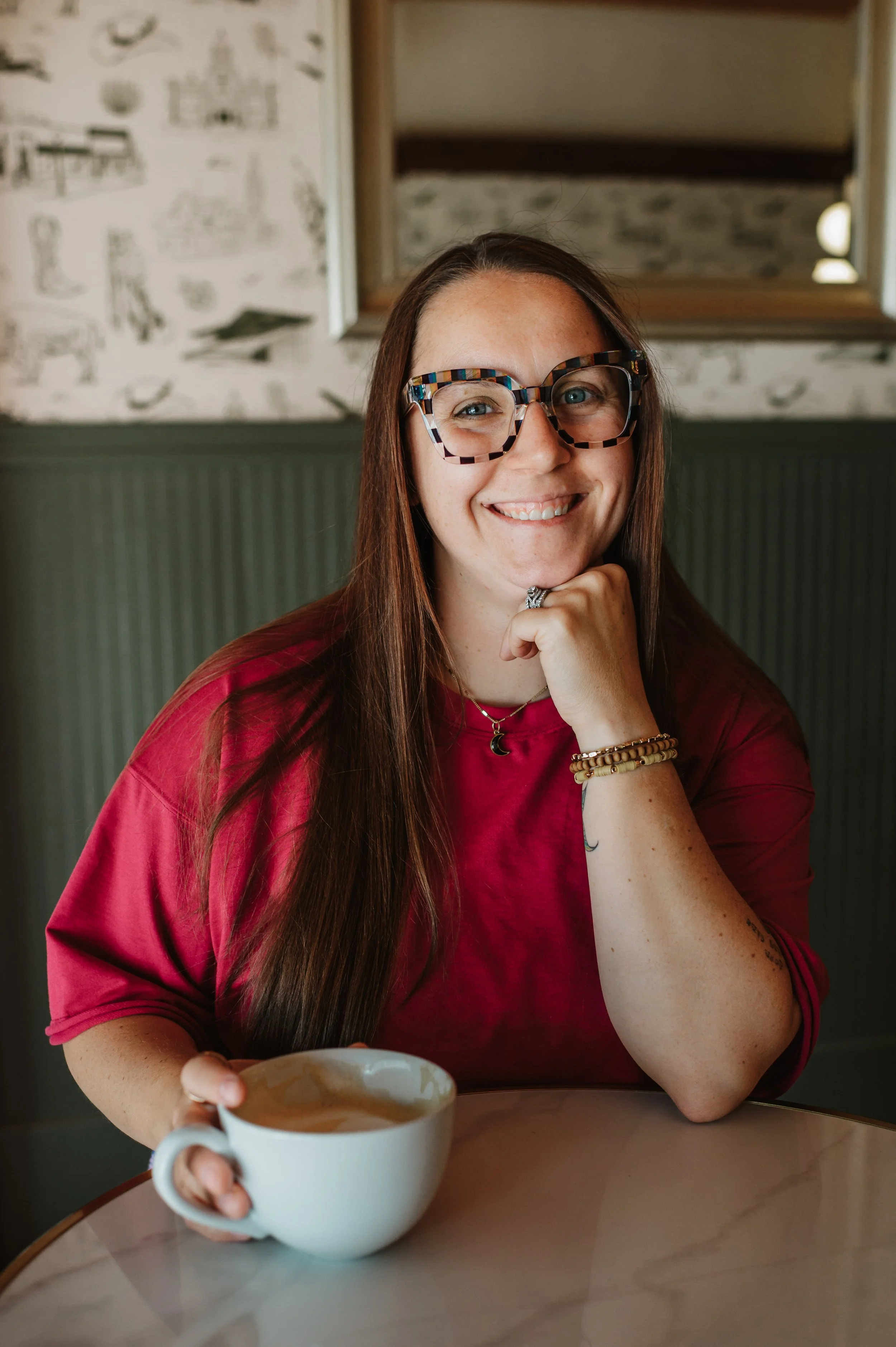 A woman with long brown hair wearing colorful glasses, a red shirt, and jewelry, smiling, holding a white mug with a hot beverage, sitting at a table in a cozy cafe or restaurant.