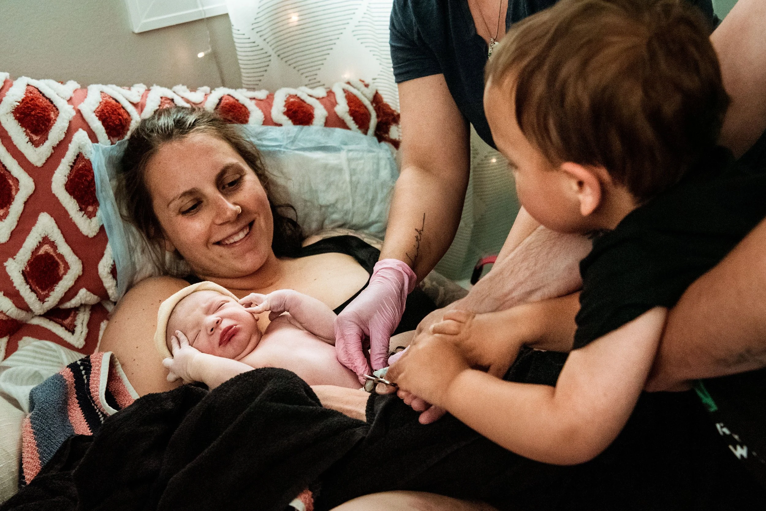 A woman with a big smile, lying in bed, is holding a newborn baby while a young boy watches, touching the baby's hand.