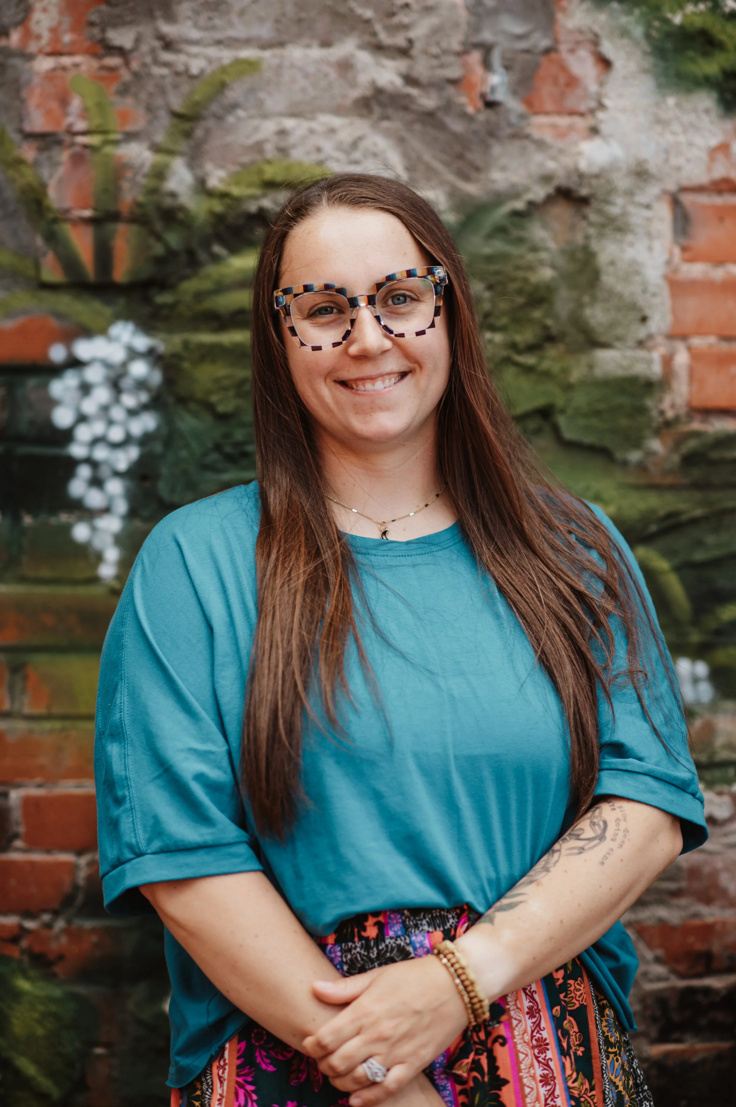 A woman with long brown hair and glasses with a colorful frames, wearing a blue top, standing in front of a weathered brick and mossy wall, smiling and looking at the camera.