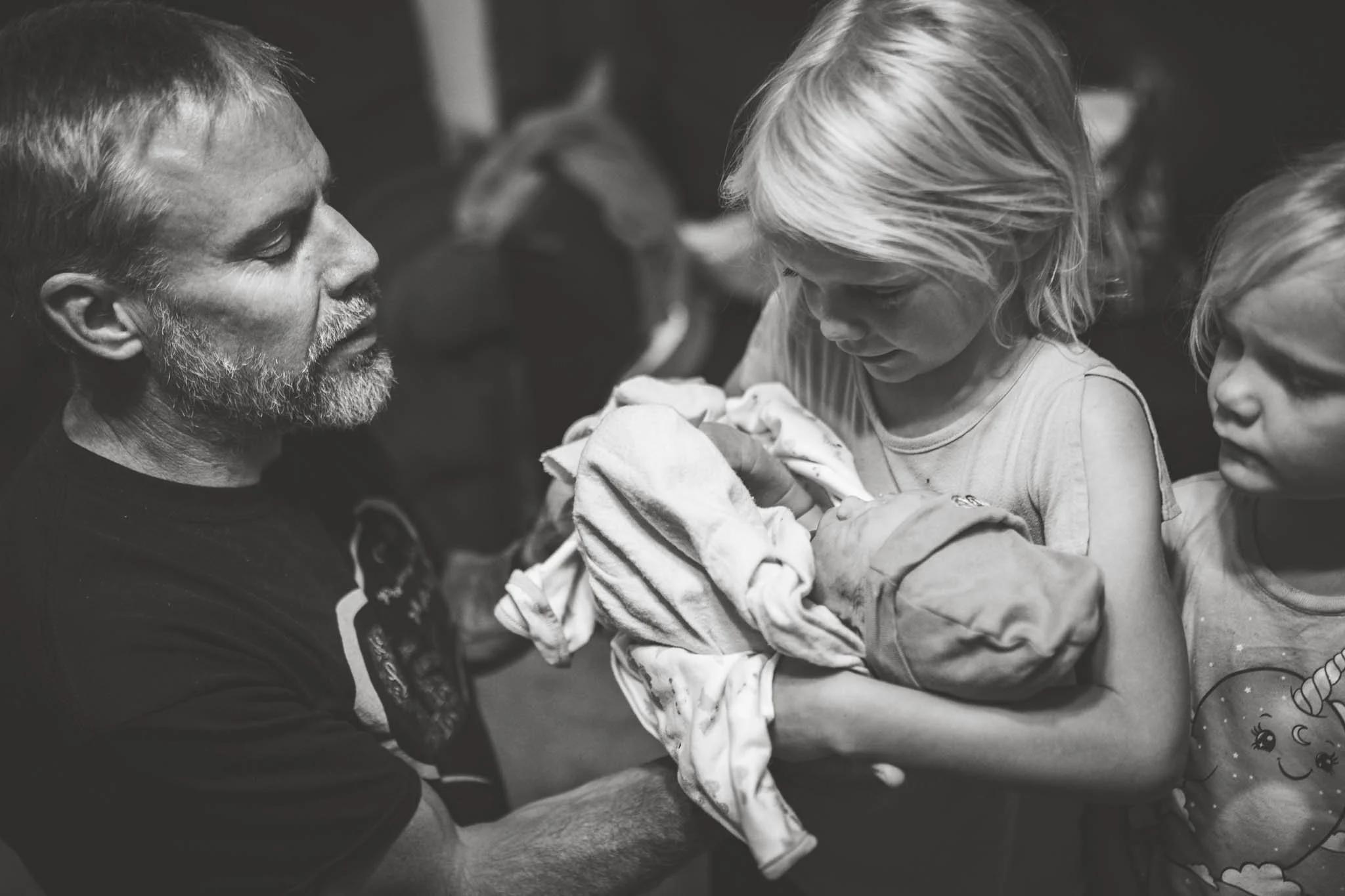 A man with a beard holding a newborn wrapped in blankets, while a young girl and another child look on closely in a black-and-white photo.