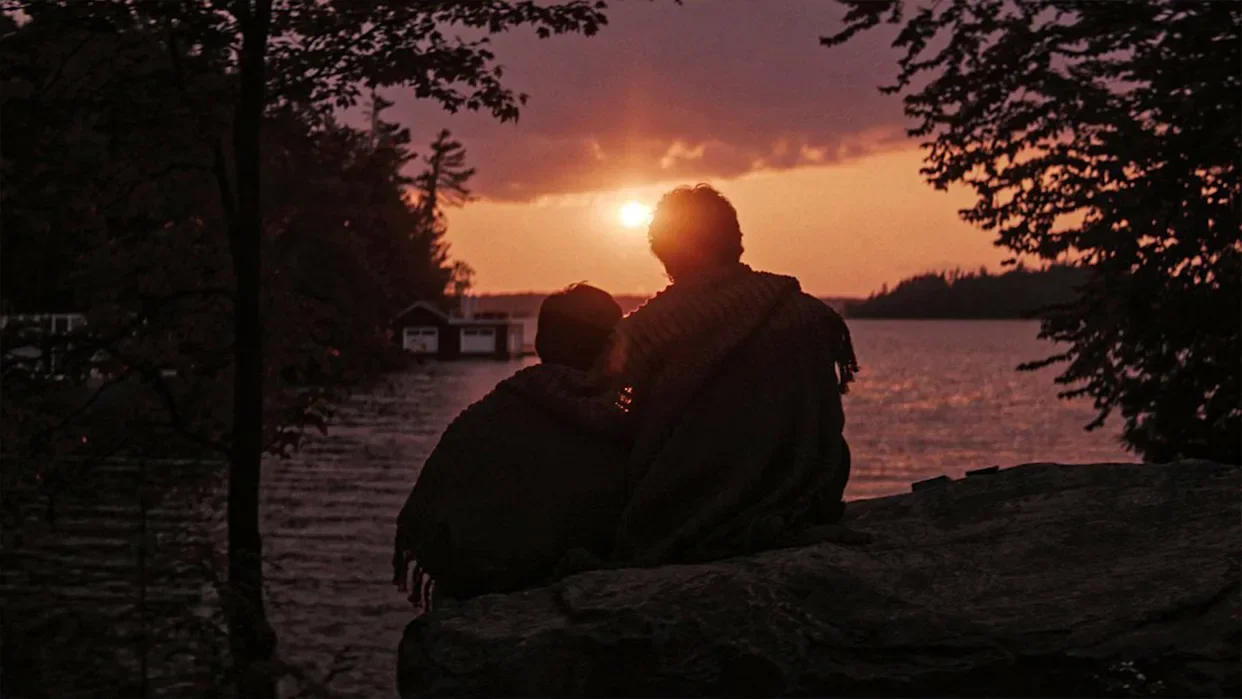 Two people sitting on rocks by a river during sunset, with trees on either side and a cloudy sky.