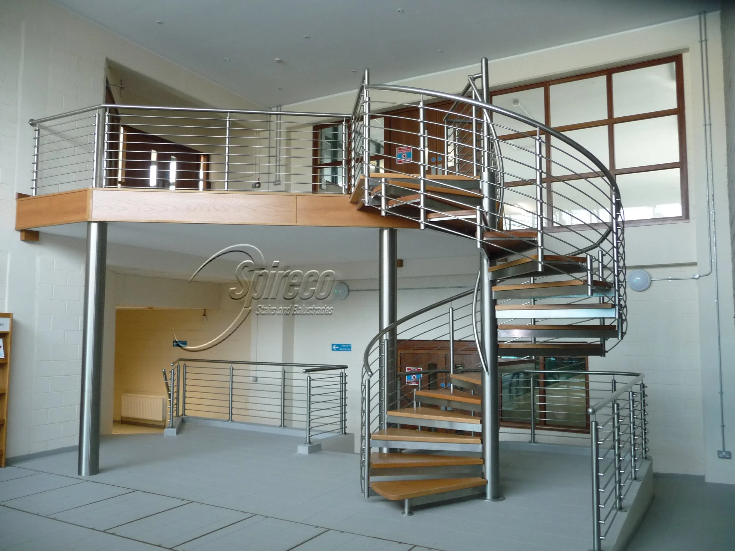 ‘Flute’ Spiral Stairs. Polished fully Stainless Steel Stairs with 2″ Red Oak treads and Landing Deck