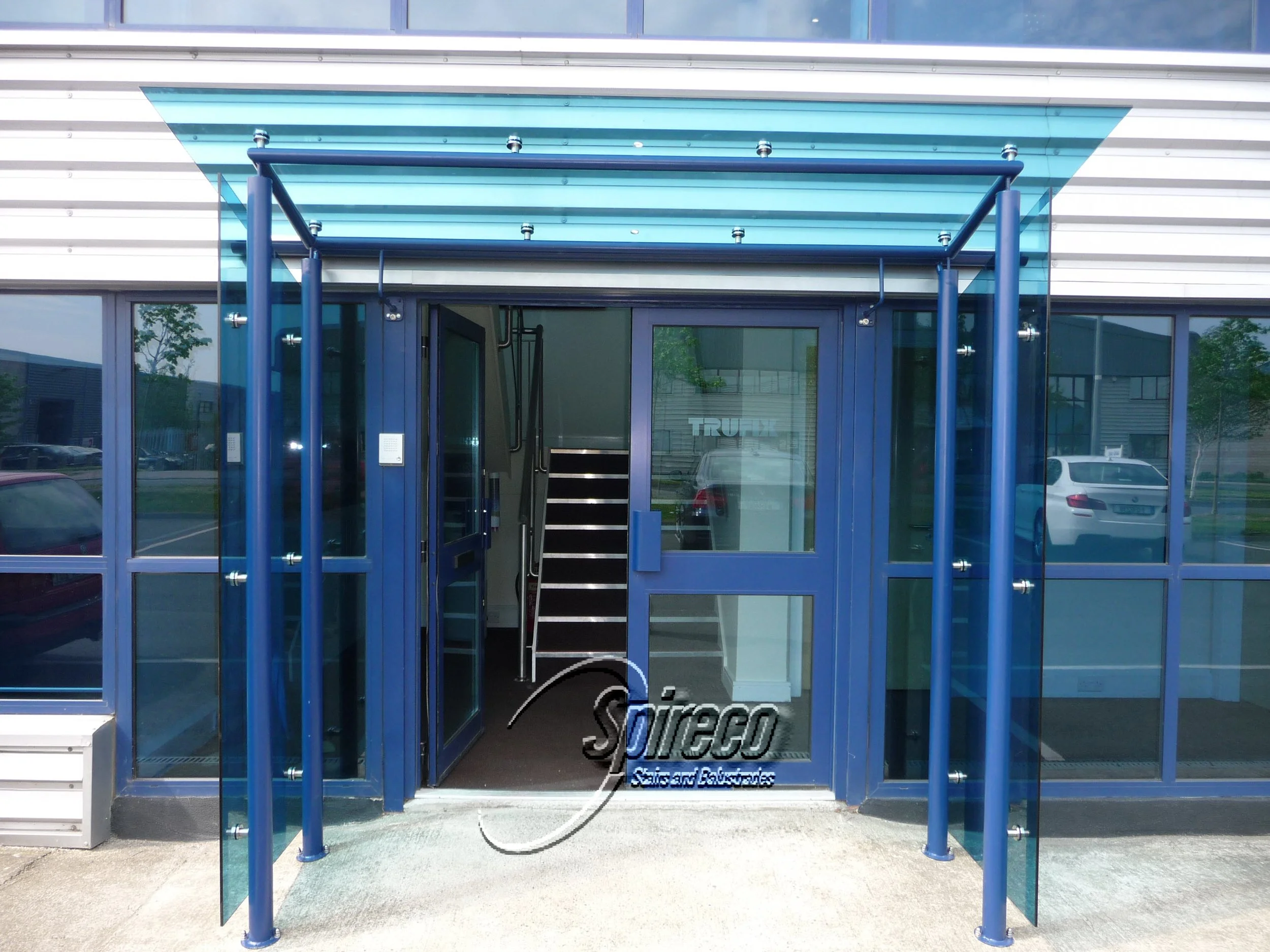 ‘Trufix’ Blue Glass Canopy.
15 mm Blue Toughened Glass connect by Stainless Steel Disc ‘Thru Clamps’ to Galvanised & Powder-coated Canopy frame. Rear Gutter collects Rainwater and channels to ground inside rear Posts.