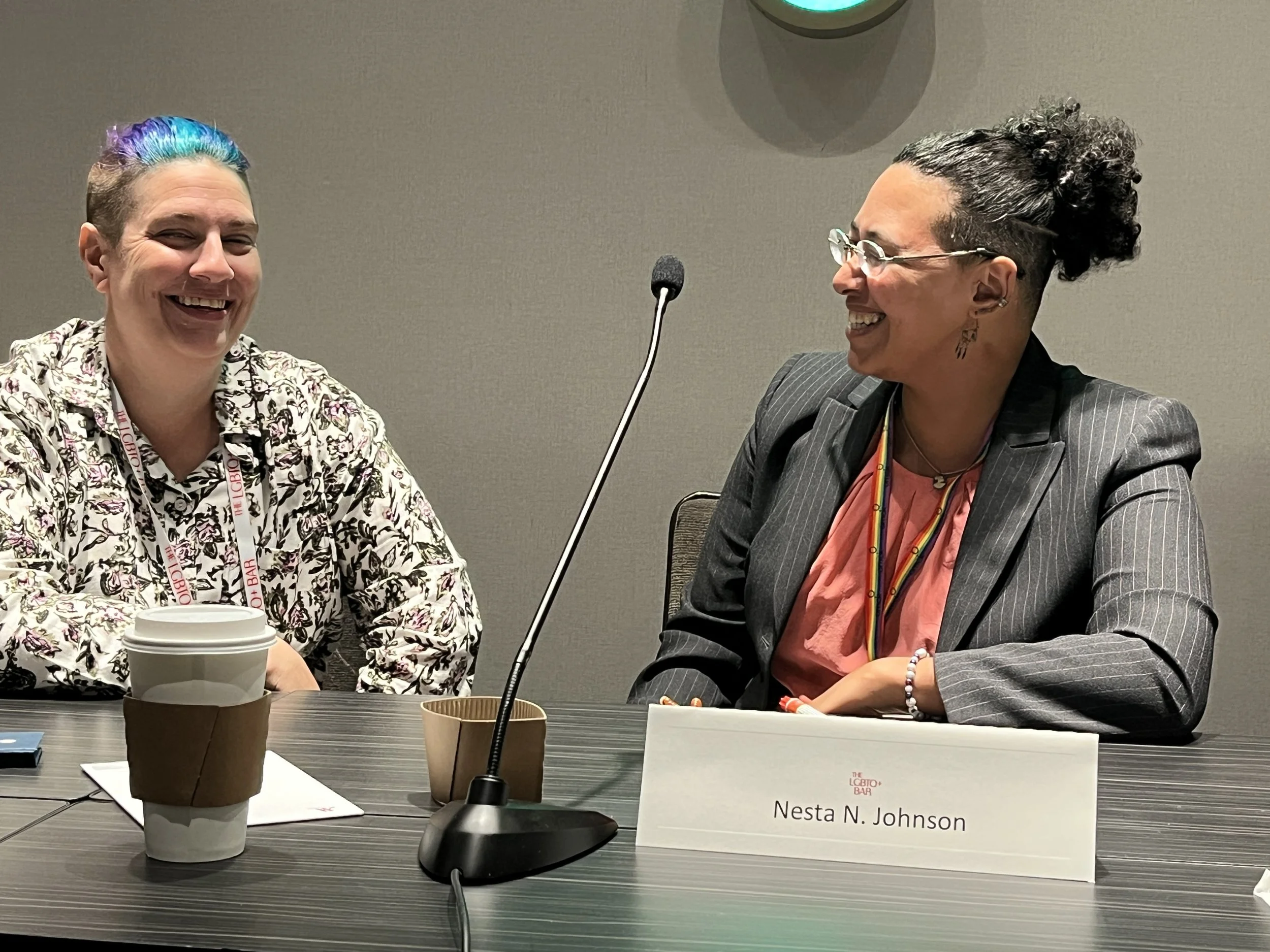 Nesta N. Johnson Rose and one other person sit at a table with microphones and name cards. Both people are laughing. Nesta's hair is up; she is wearing glasses, a salmon-pink blouse, and a grey pinstriped blazer. Photo taken at Lavender Law Conferenc