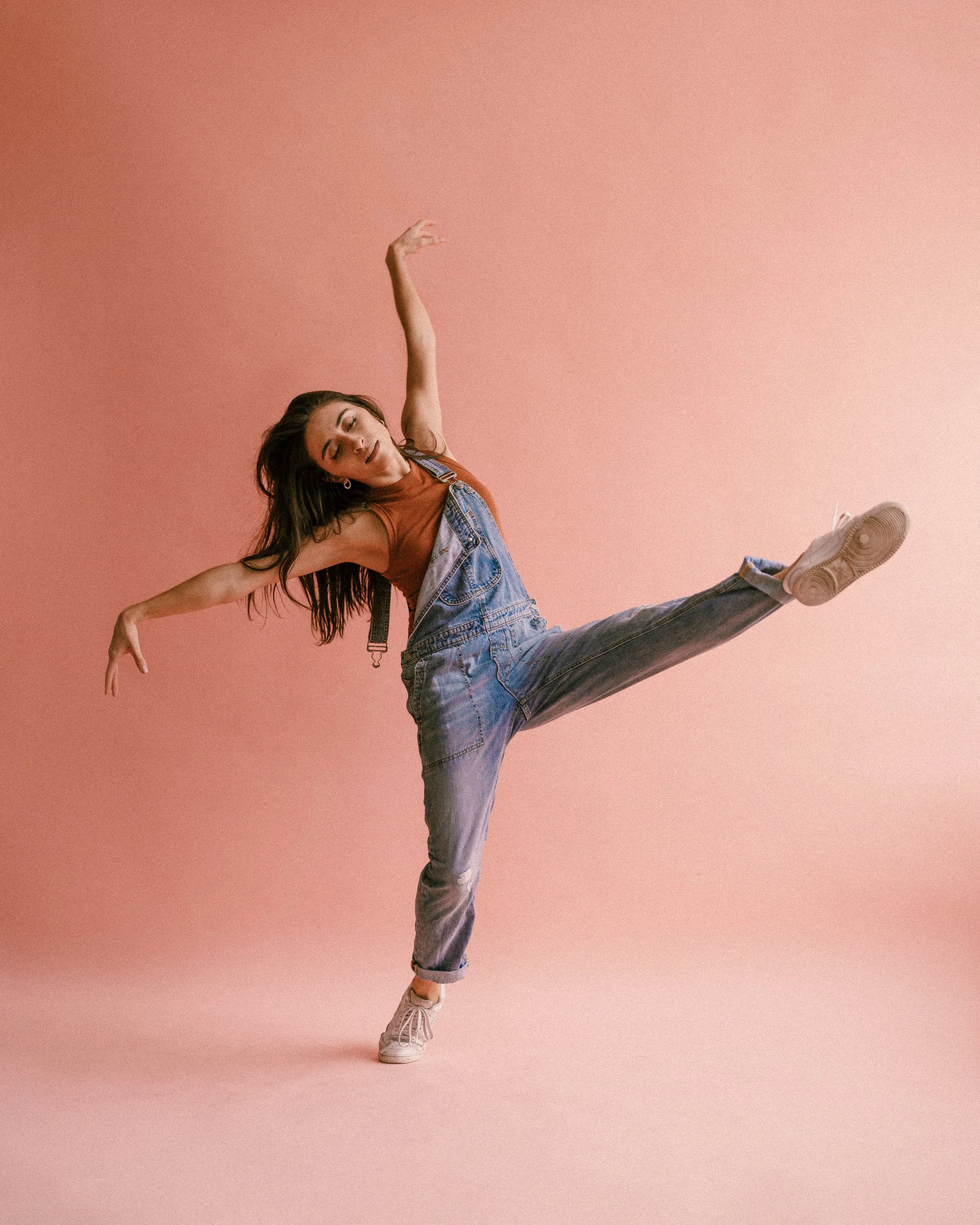 A woman dancing against a pink background, wearing denim overalls and a brown top, balancing on one leg with arms raised and eyes closed.