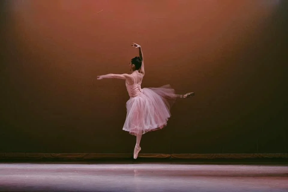 A ballet dancer in a pink tutu performing a leap during a dance recital on stage.