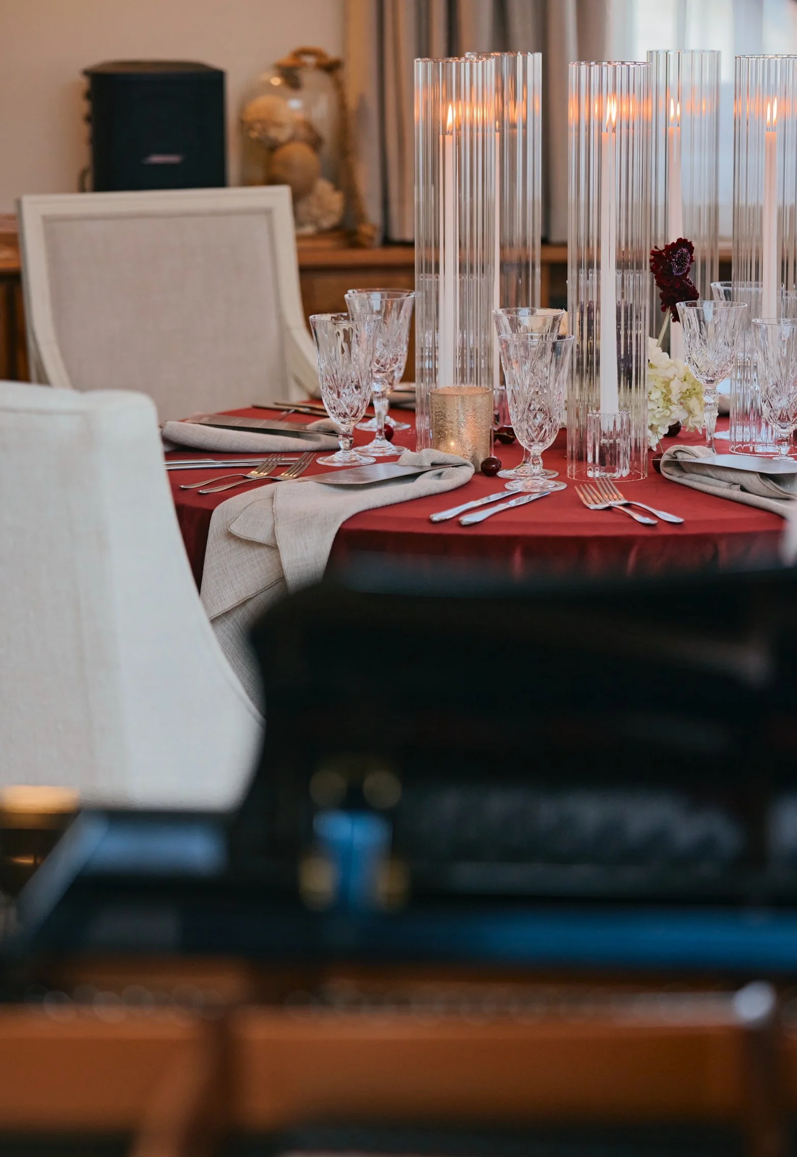 Table élégante avec vaisselle en cristal, bougies, fleurs blanches et rouge profond, nappes rouges et serviettes gris clair, décor intérieur chaleureux.