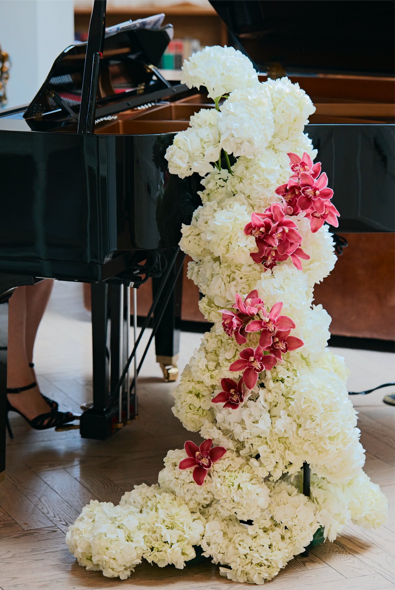 Un piano noir décoré avec un arrangement de fleurs blanches et roses, comprenant des hydrangeas et des orchids, dans un intérieur probable d'une salle de concert ou d'un lieu de performance.