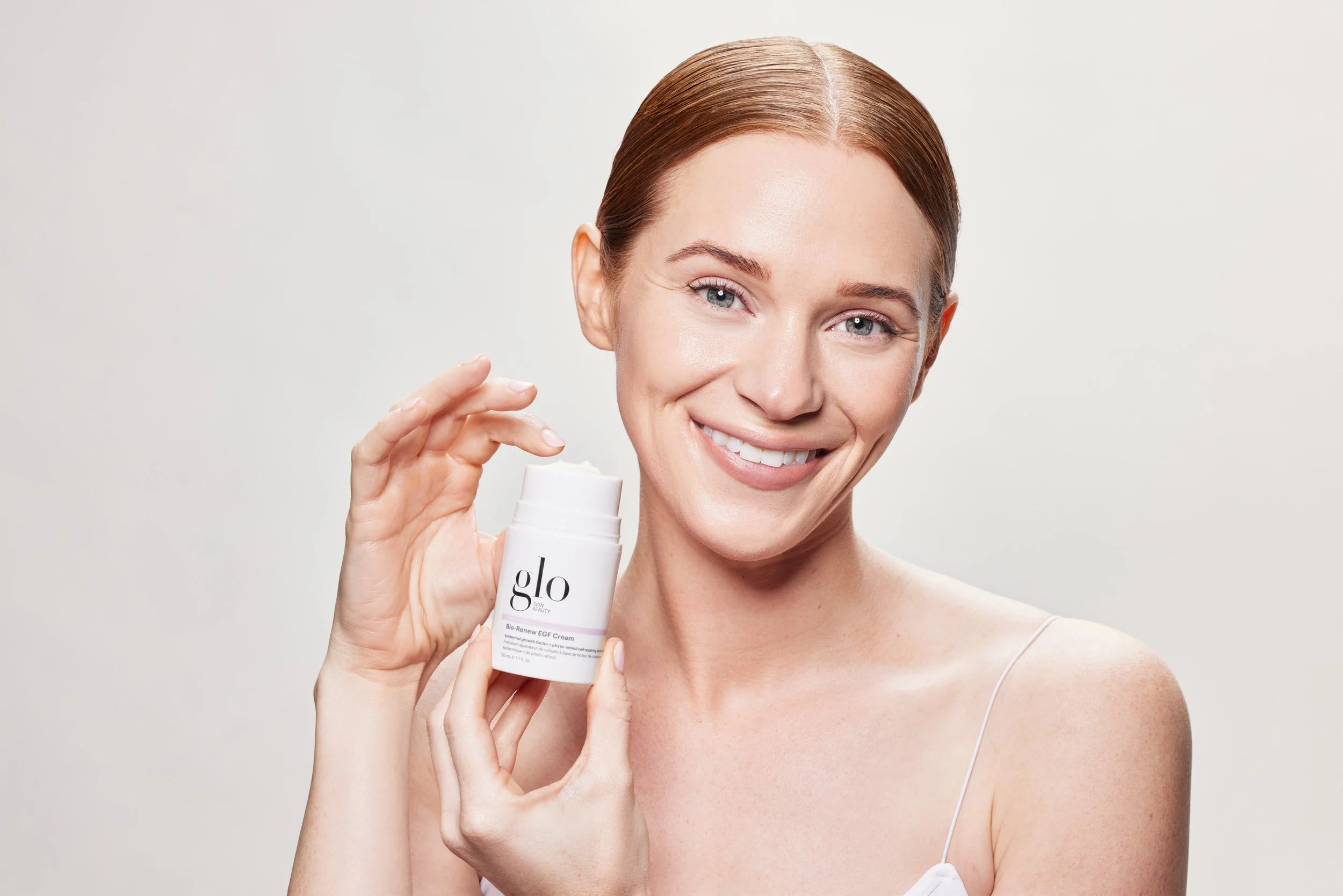 A smiling woman with red hair holding a white container of Glo Bio Renew EGF Cream.