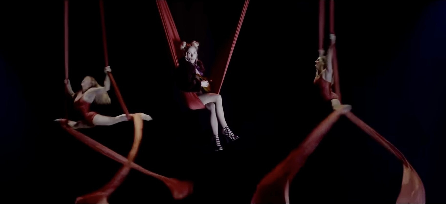 Three women on red aerial silk swings against a dark background, with the woman in the center holding a guitar.