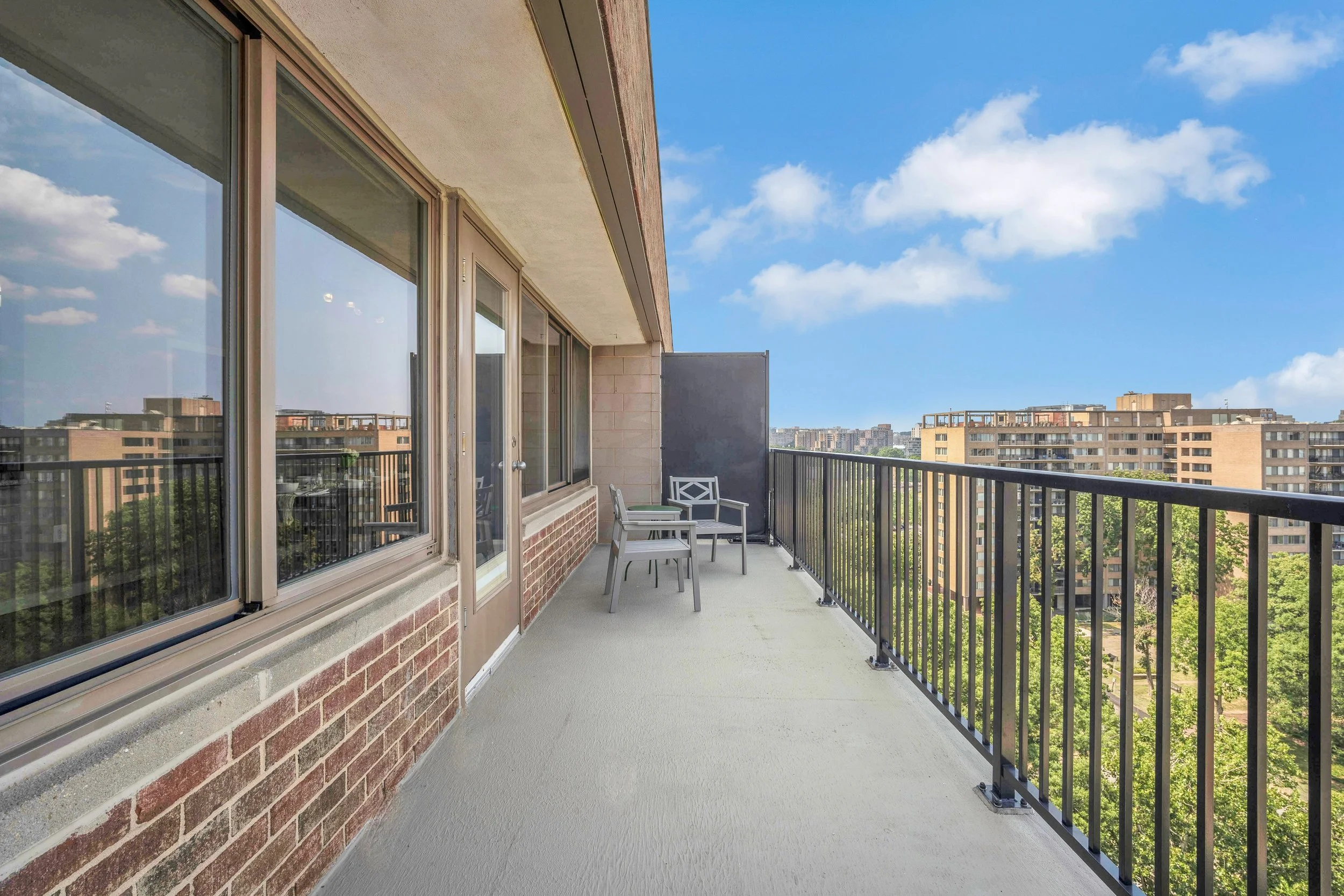Balcony with outdoor chairs and table, overlooking a city skyline under a partly cloudy blue sky.