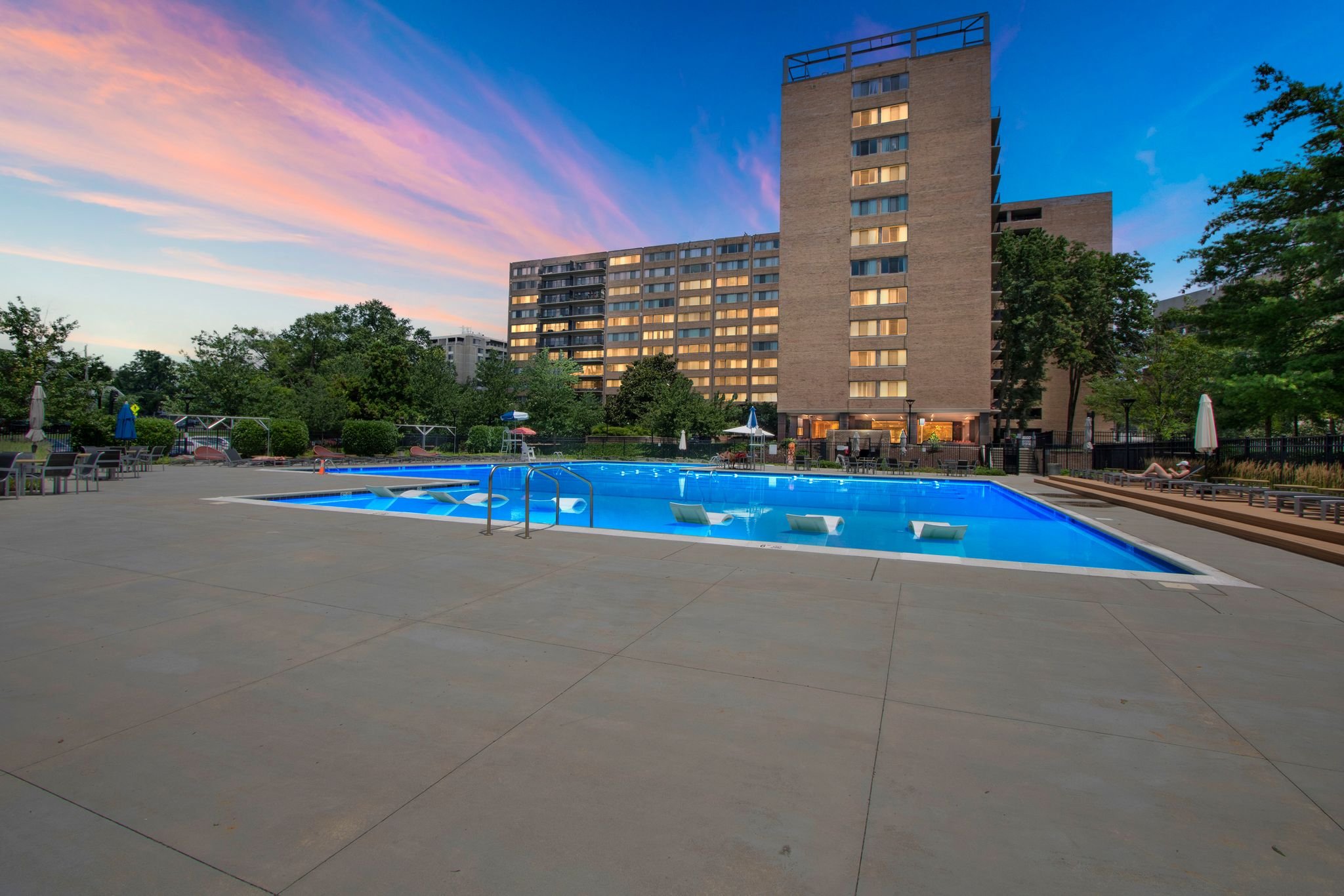 An outdoor swimming pool at sunset, surrounded by lounge chairs and umbrellas, with tall apartment buildings and green trees in the background.