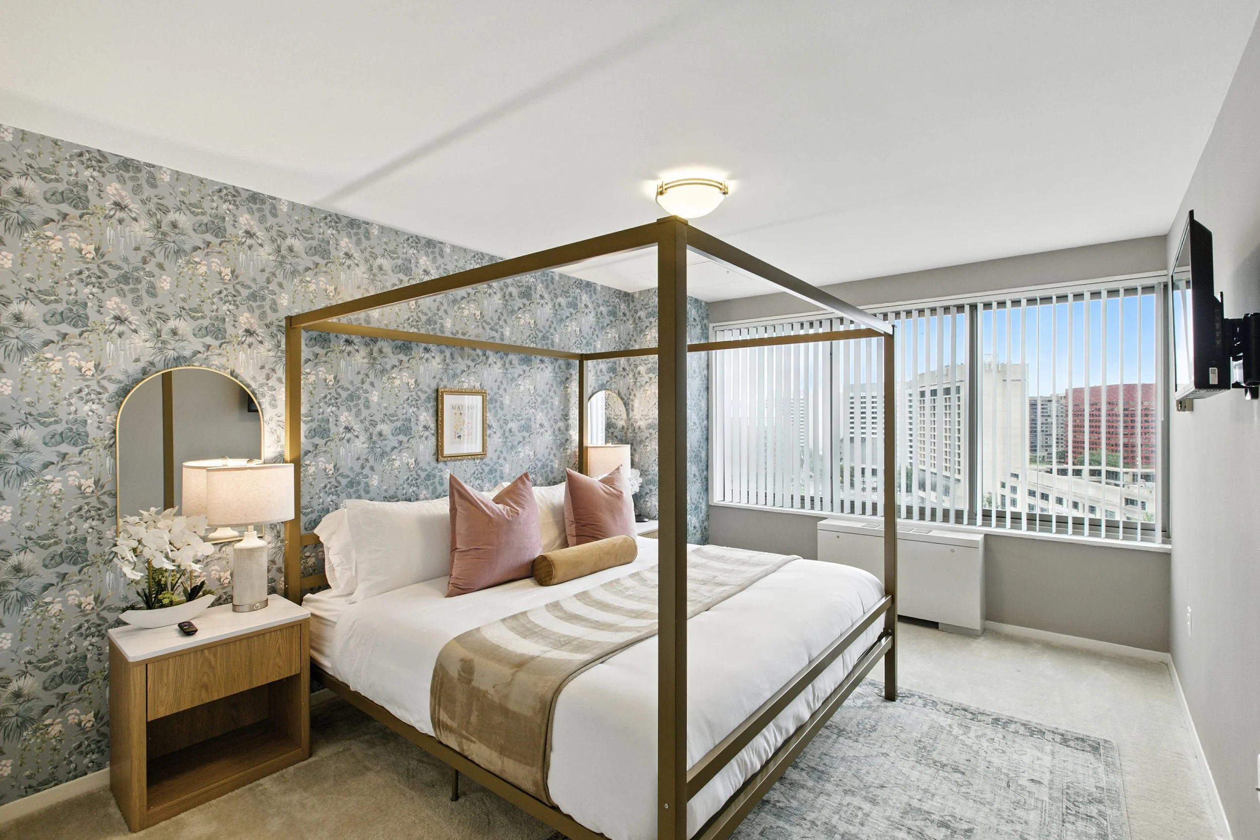 A modern bedroom with a four-poster bed, pink pillows, a beige and white striped bed runner, and a floral accent wall. Has a nightstand with a lamp and flowers, and large windows with vertical blinds showing city buildings outside.