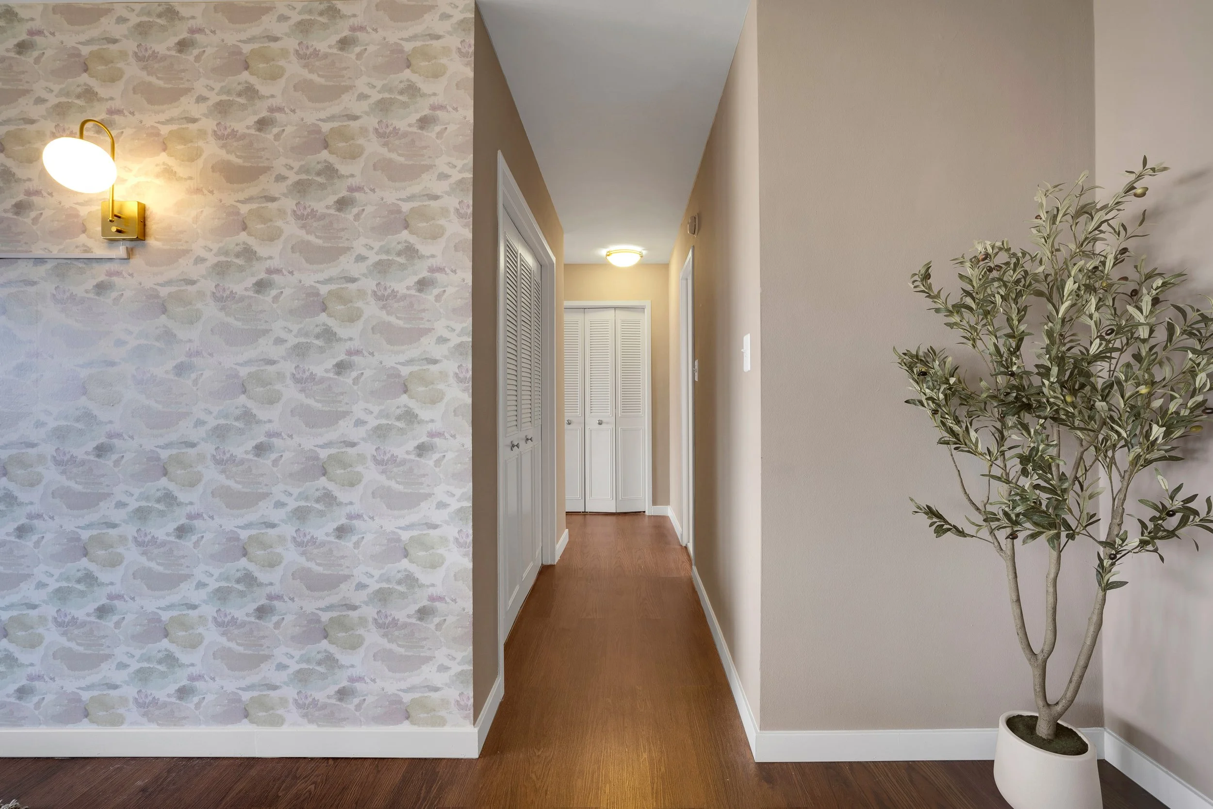 Empty hallway with wooden flooring, beige walls, a wall-mounted light with a white shade on the left, and a potted plant on the right. Closet doors are visible at the end of the hallway.