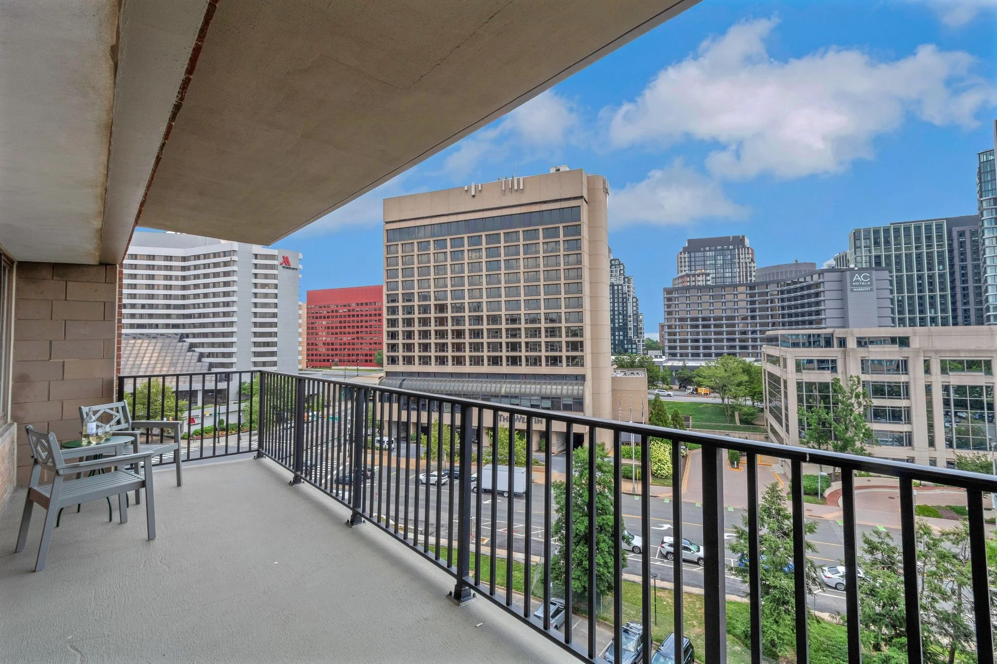 Balcony with two chairs and a small table overlooking a cityscape with tall buildings, trees, and a parking lot on a sunny day with blue sky and some clouds.