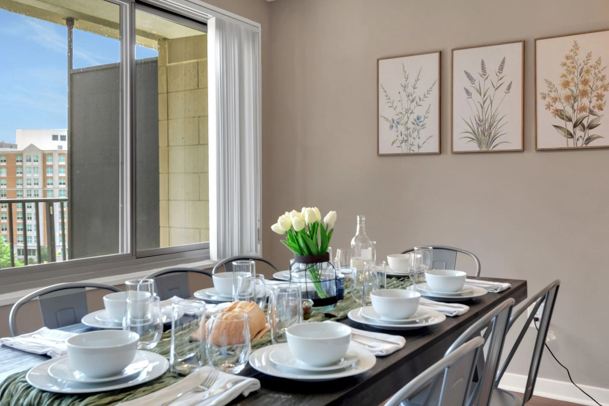 A dining table set with white bowls, glasses, and silverware, with a vase of white tulips in the center, in a room with a large window and framed botanical artwork on the wall.