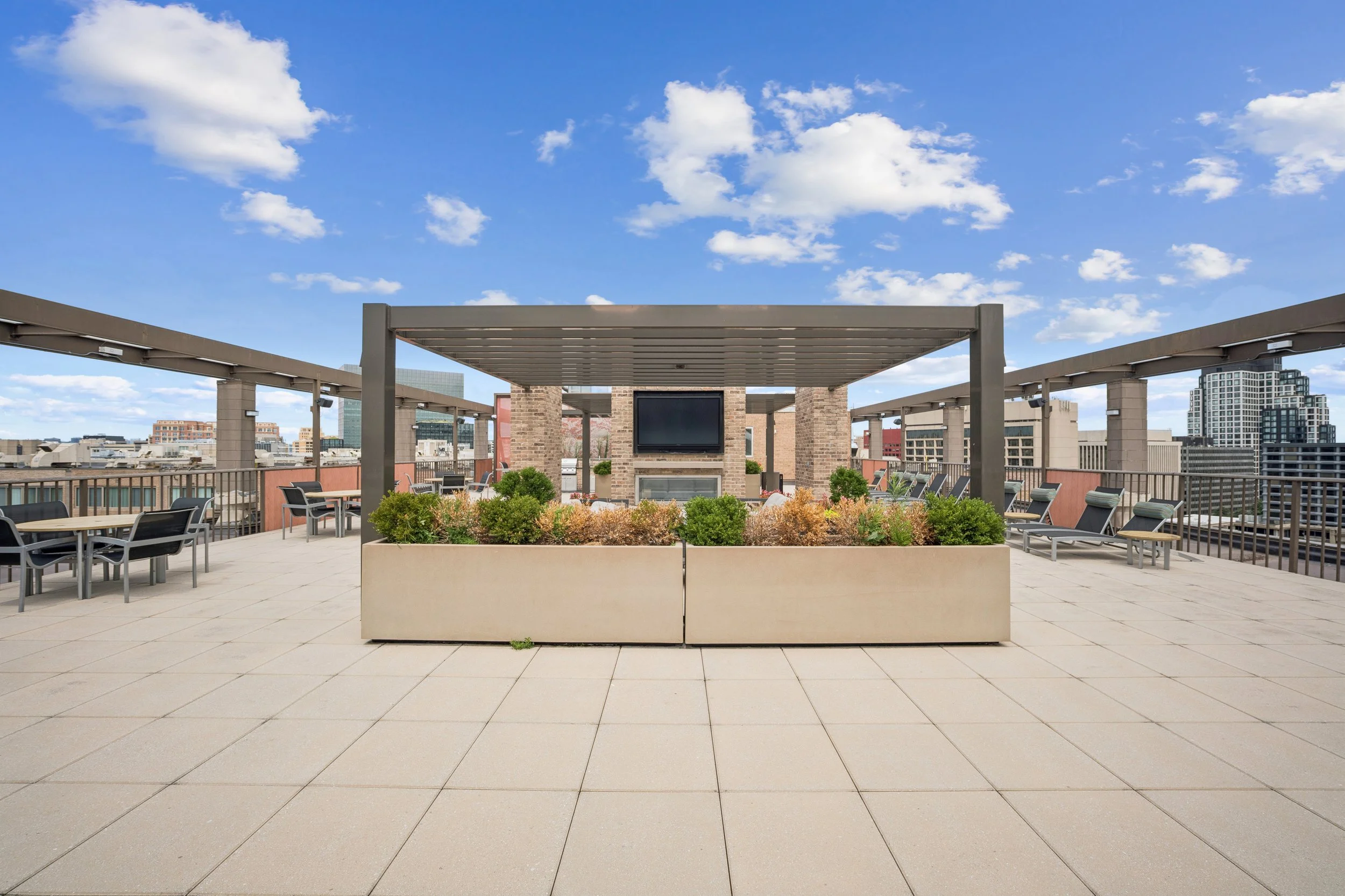 Rooftop terrace with seating area and a large flat-screen TV over a fireplace, surrounded by potted plants and city skyline skyline under a blue sky with white clouds.
