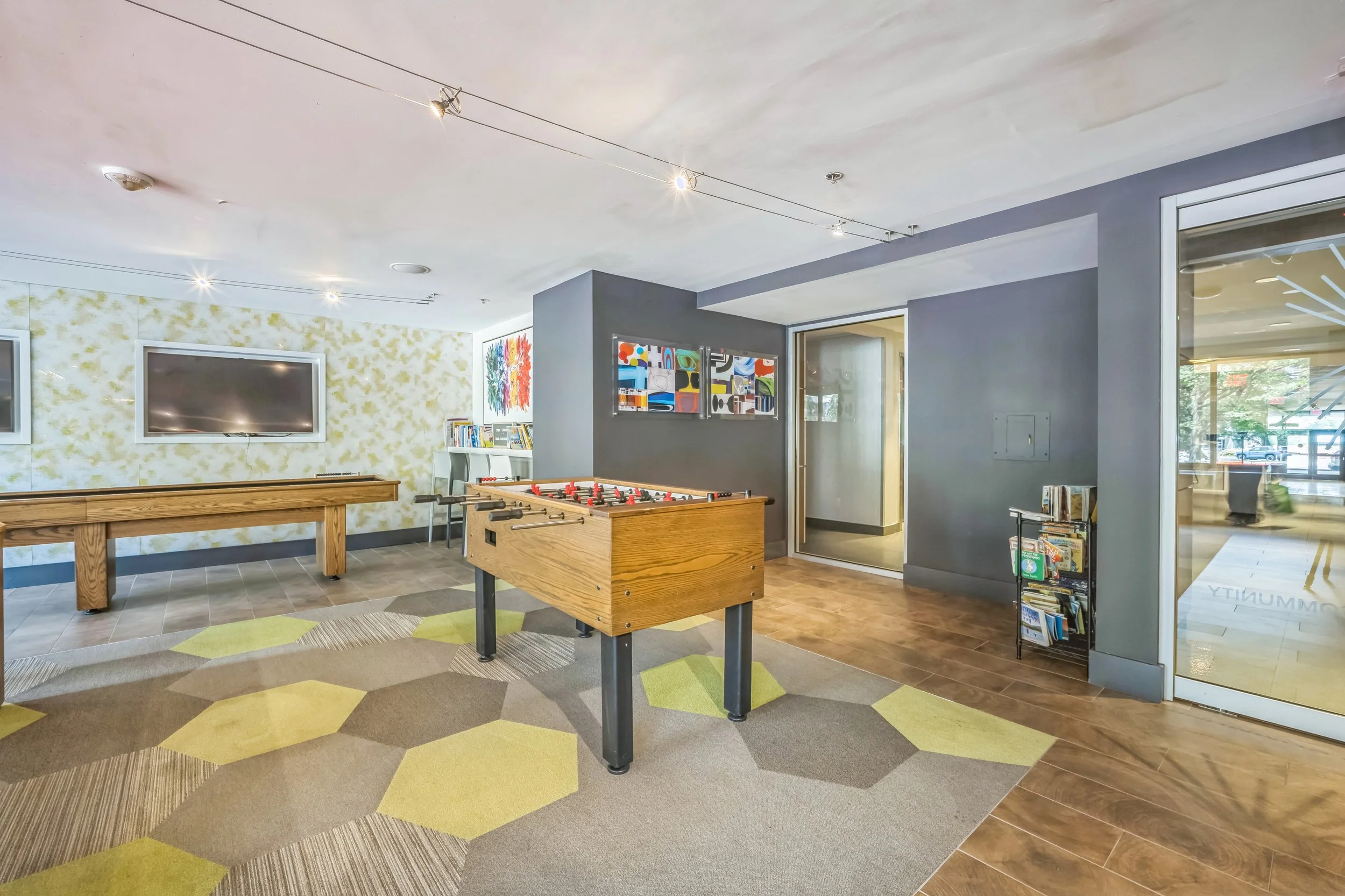 Indoor recreational room with a foosball table, wall-mounted televisions, colorful artwork, bookshelves, a large window, and a carpet with yellow and green hexagon patterns.