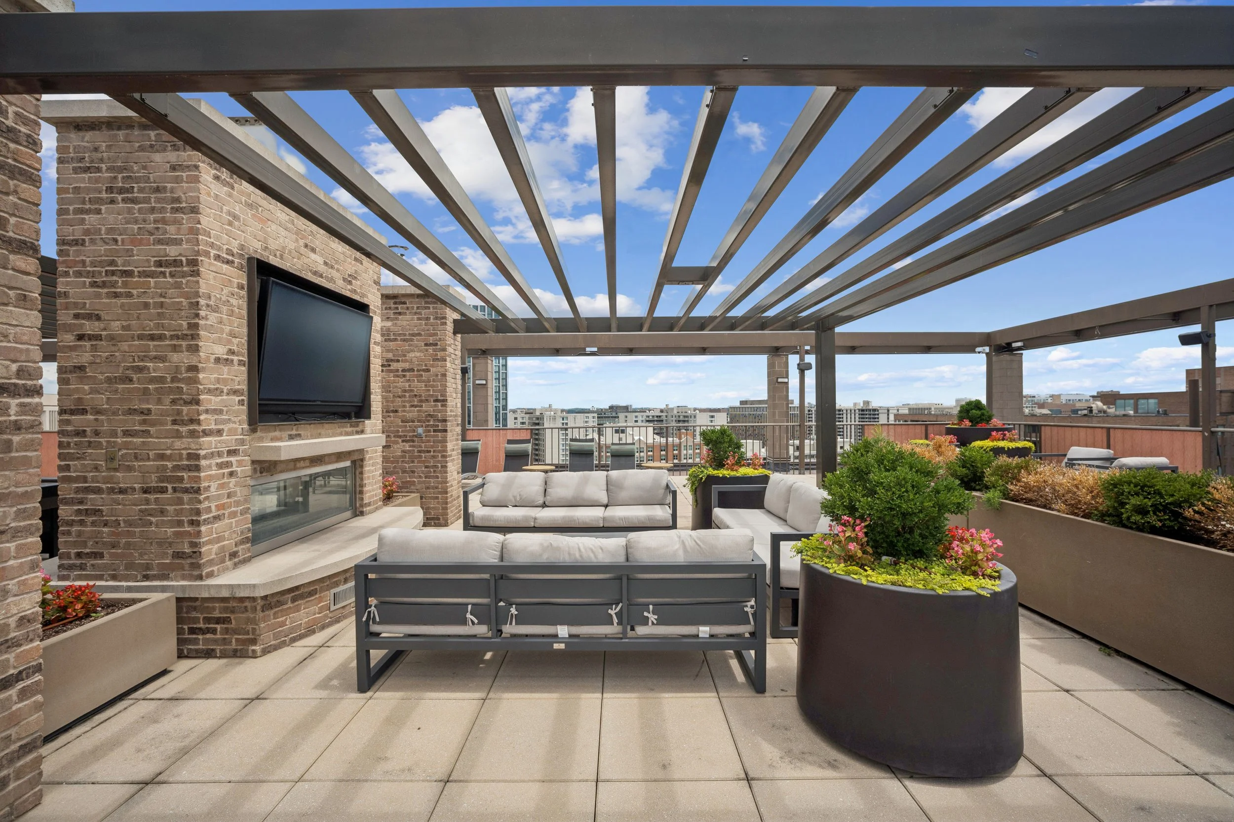 Rooftop patio with brick walls, outdoor seating, planters with greenery, and city skyline under a partly cloudy sky.