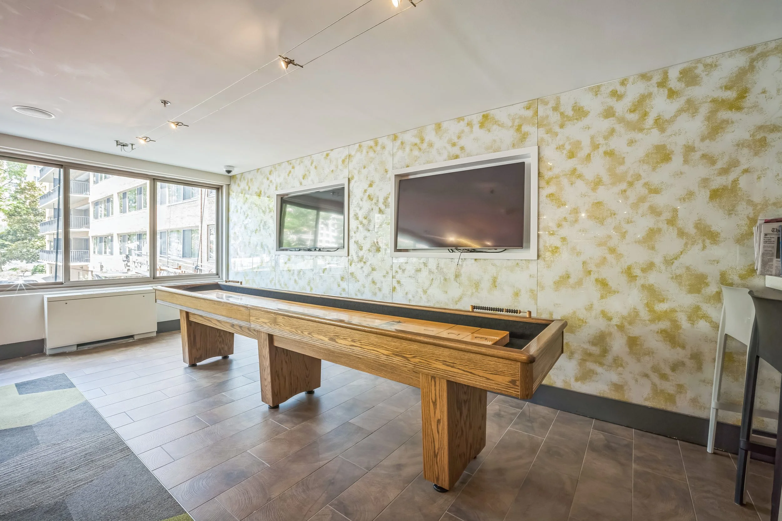 Indoor recreation room with a shuffleboard table, two wall-mounted TVs, large windows, and modern decor.