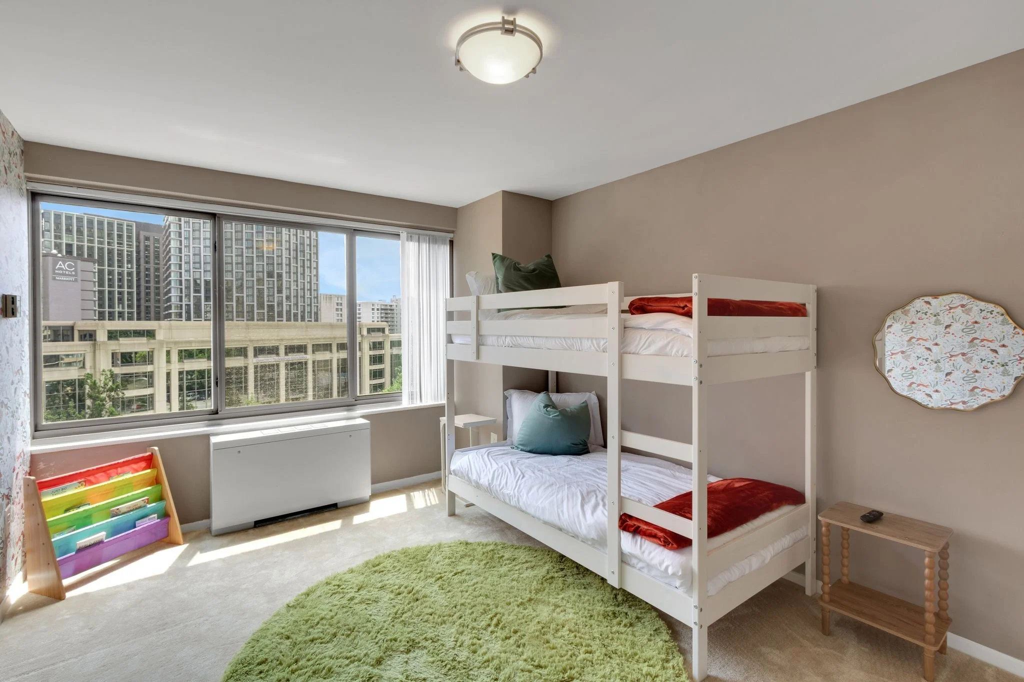 A bedroom with a bunk bed, a green round rug, a nightstand with a remote, a mirrored wall piece, and a large window showing city buildings.