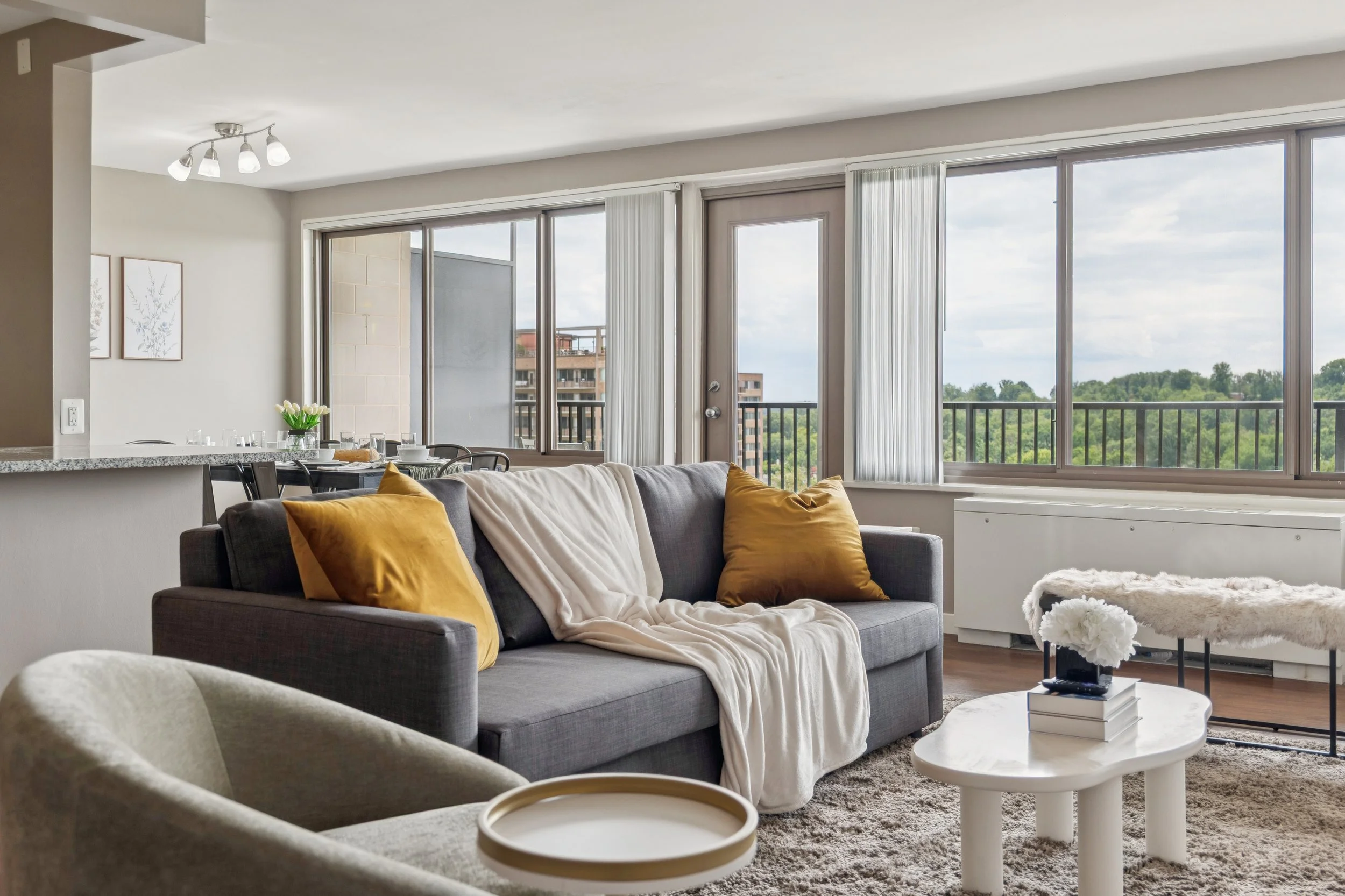 Modern living room with large windows, gray sofa with gold pillows, white blanket, white coffee table with flowers and books, beige armchair, and a view of buildings and greenery outside.
