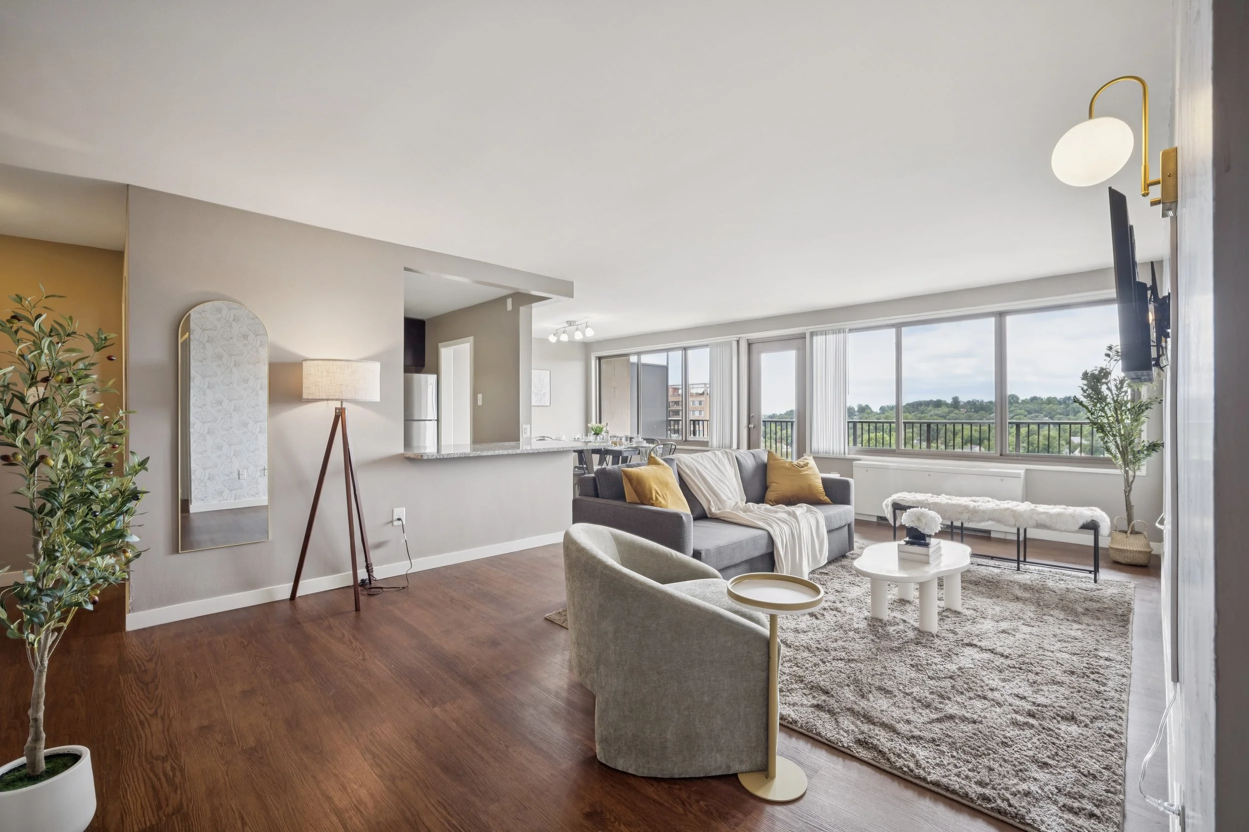 Modern living room with large windows, gray sofa, white coffee table, beige armchair, and decorative plants.