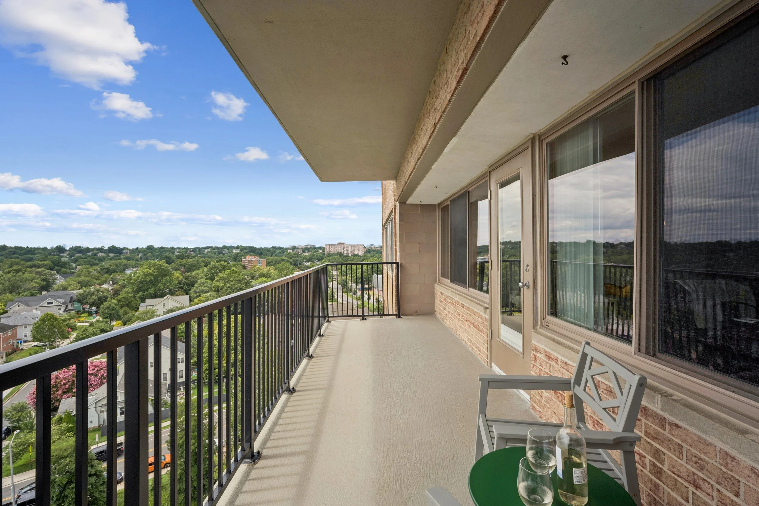 Balcony view from the apartment overlooking a green neighborhood with trees, houses, and a partly cloudy sky.