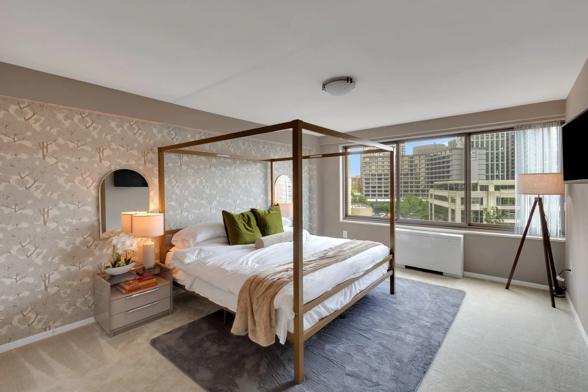 A bedroom with a four-poster bed, green pillows, and a beige throw blanket. There is a nightstand with a lamp, a bowl, and books. A window shows tall buildings and trees outside. The room has light-colored carpet and contemporary decor.